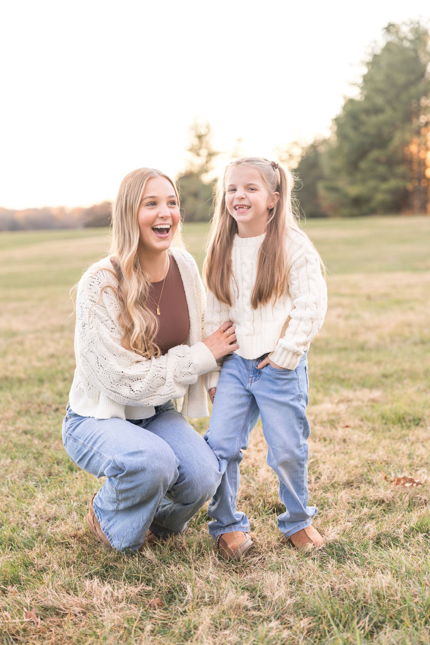Two people in white sweaters and blue jeans laugh together while sitting in a grassy field at sunset.