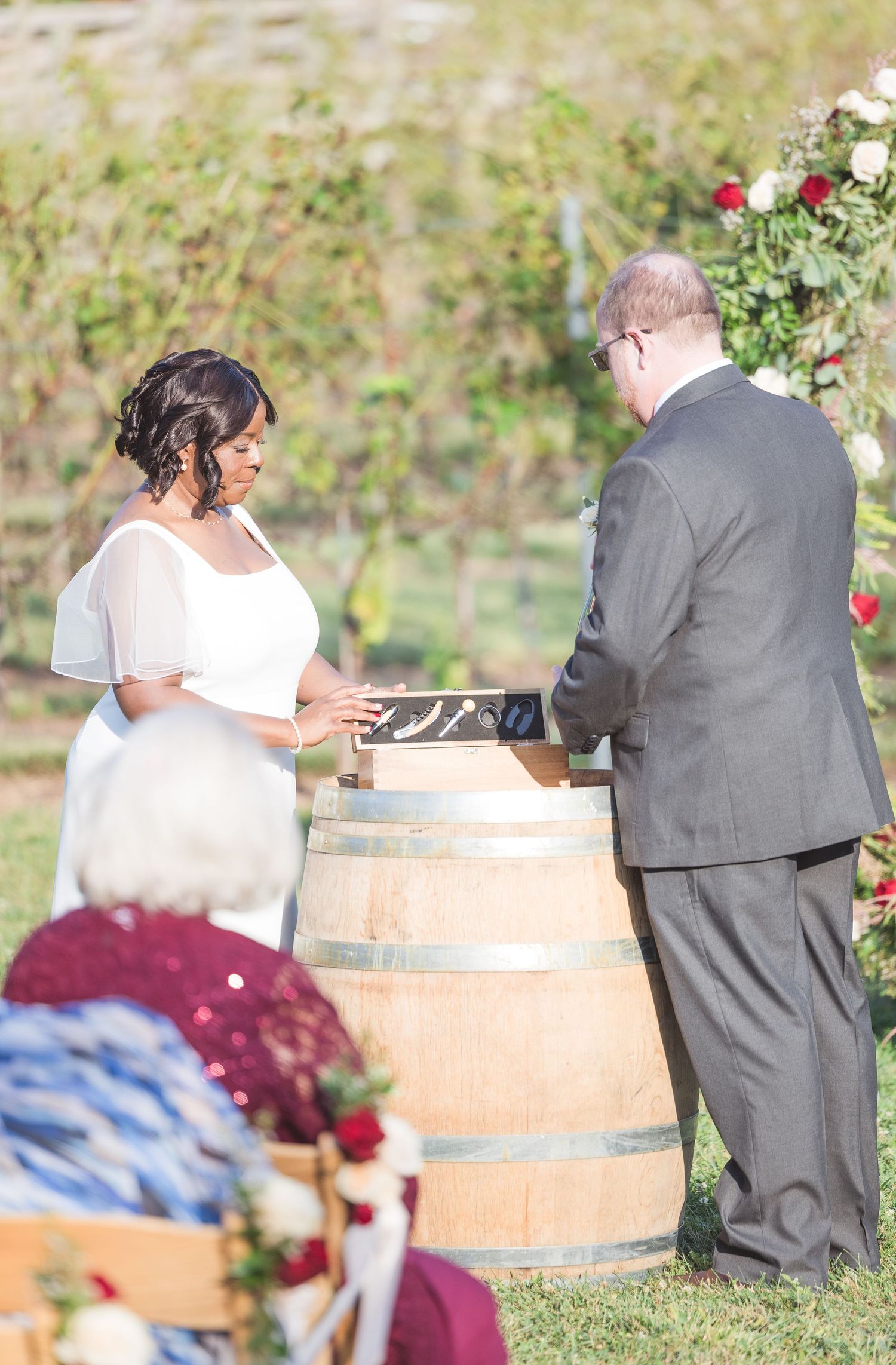 Wedding guests gather around wooden barrels during an outdoor vineyard ceremony.