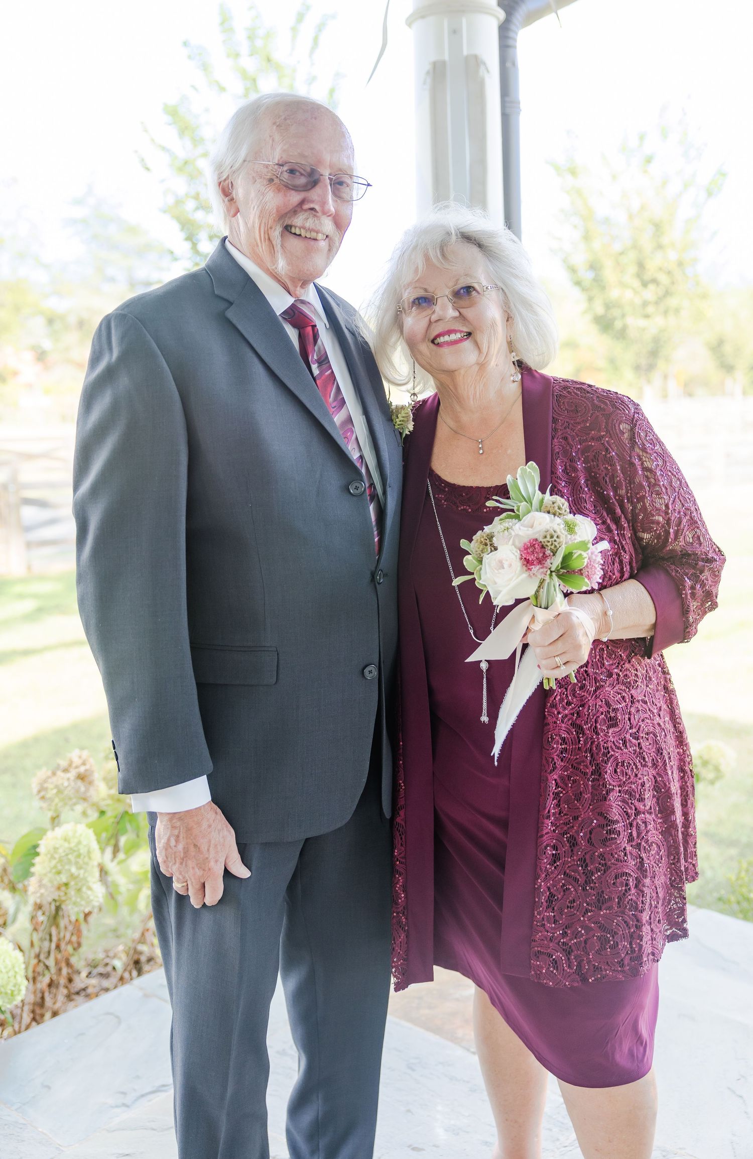An elderly couple poses together outdoors wearing formal attire in burgundy and black.
