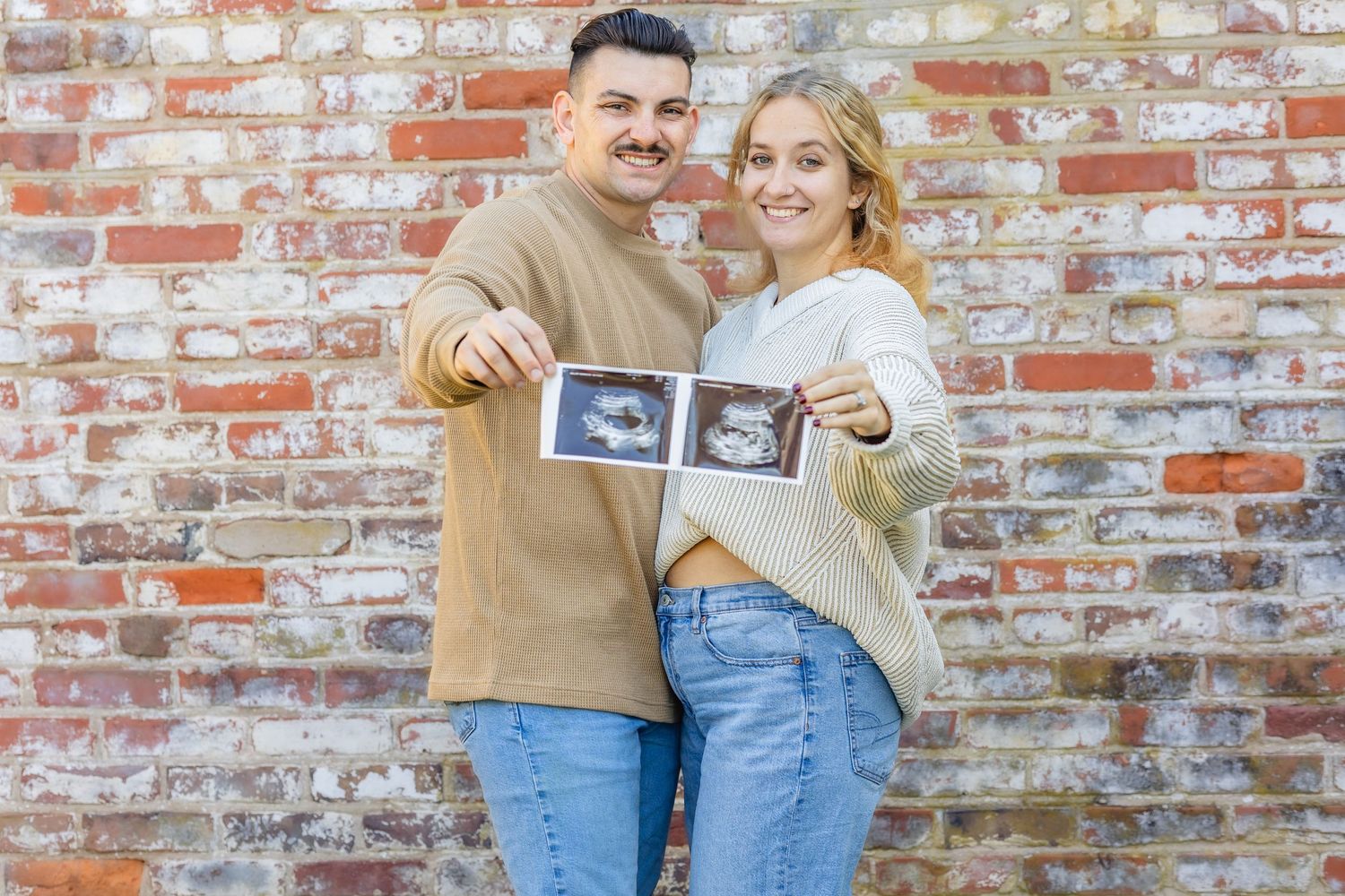 A couple poses together against a rustic brick wall holding ultrasound photos in a pregnancy announcement.