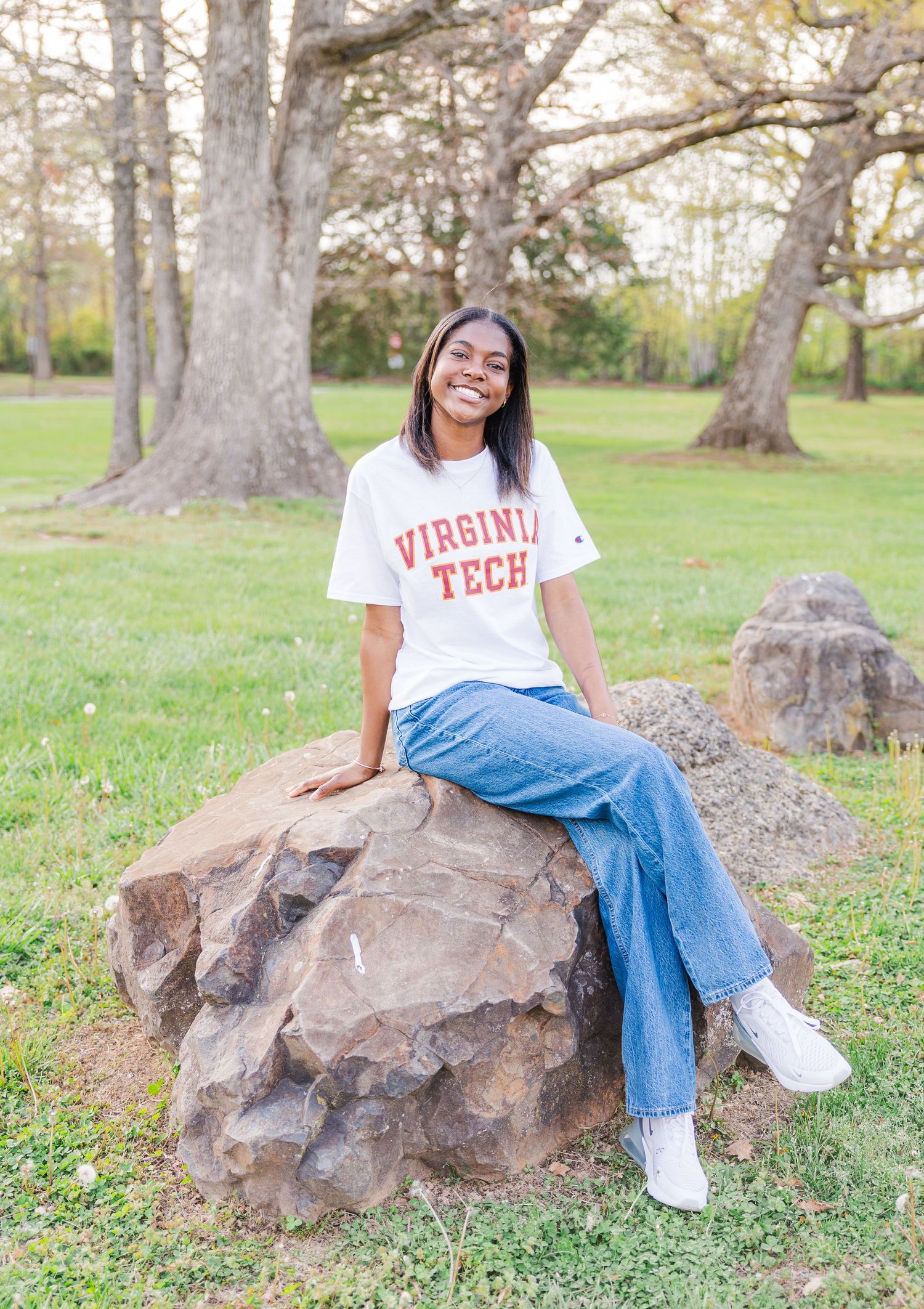 A person in university attire sits on large rocks in a sunny outdoor park setting.