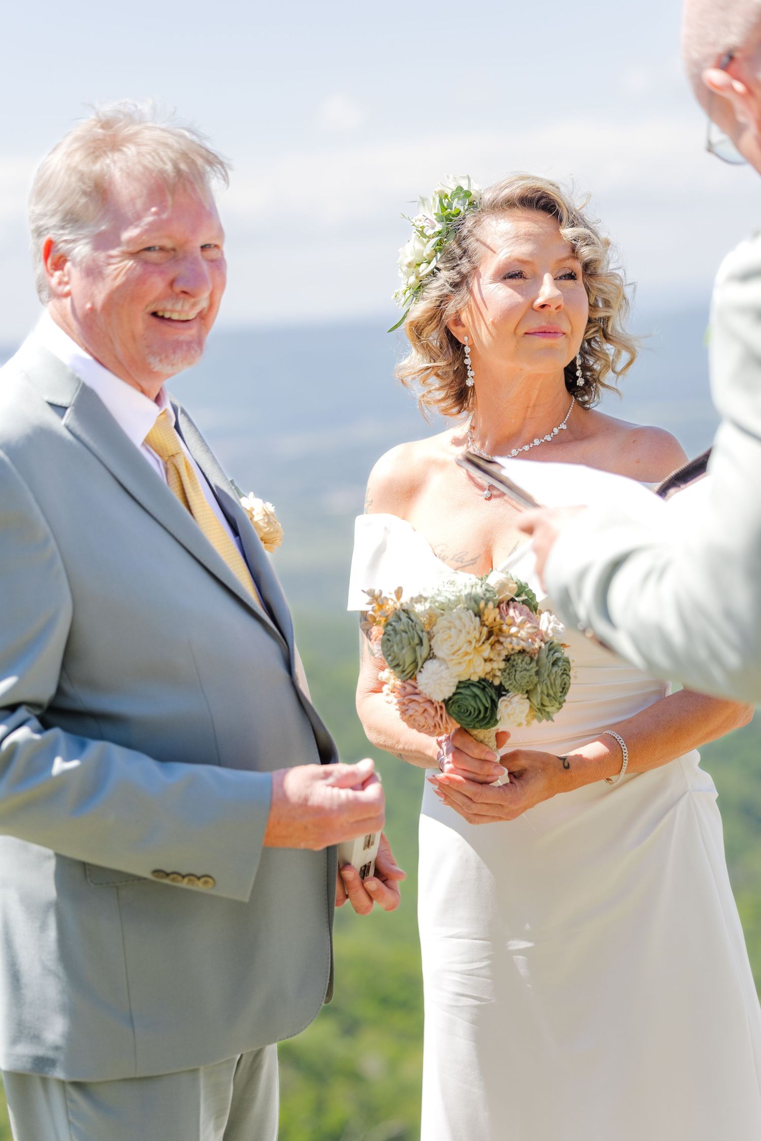 Bride holding succulent bouquet stands with groom in gray suit during outdoor mountain wedding ceremony.