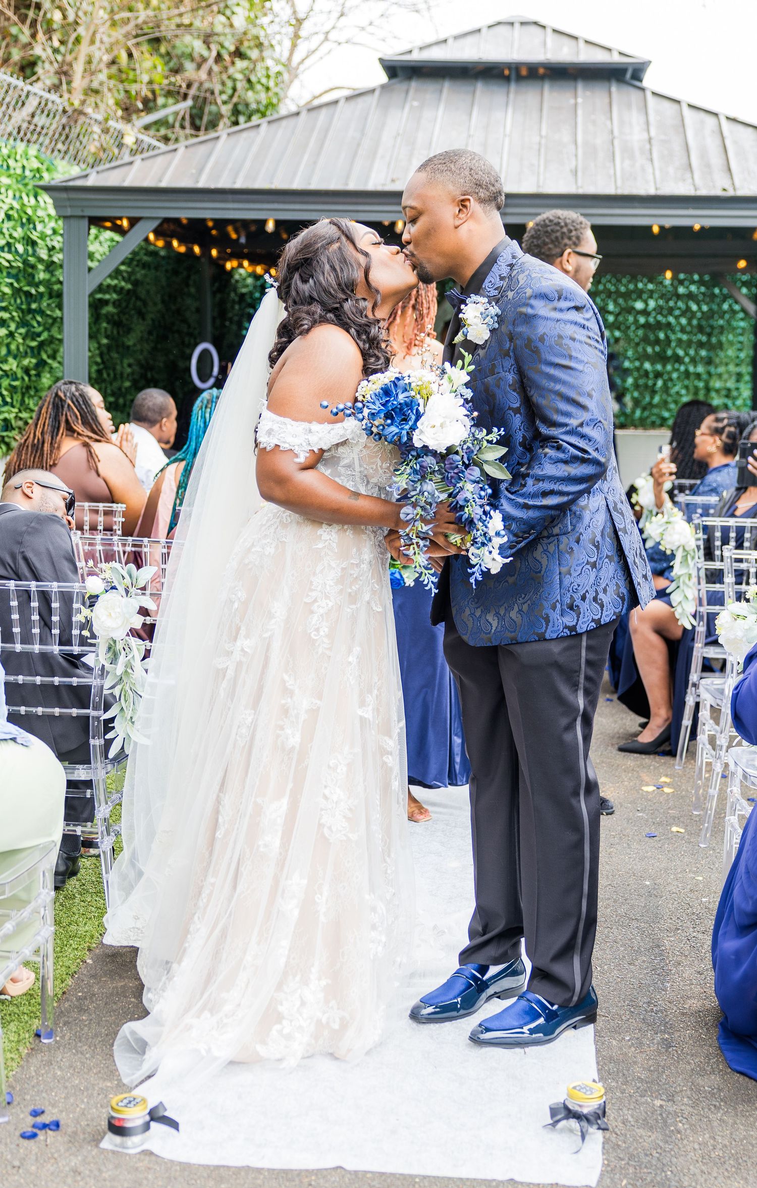Couple in navy blue and white wedding attire embrace during outdoor ceremony with white chairs and gazebo setting.