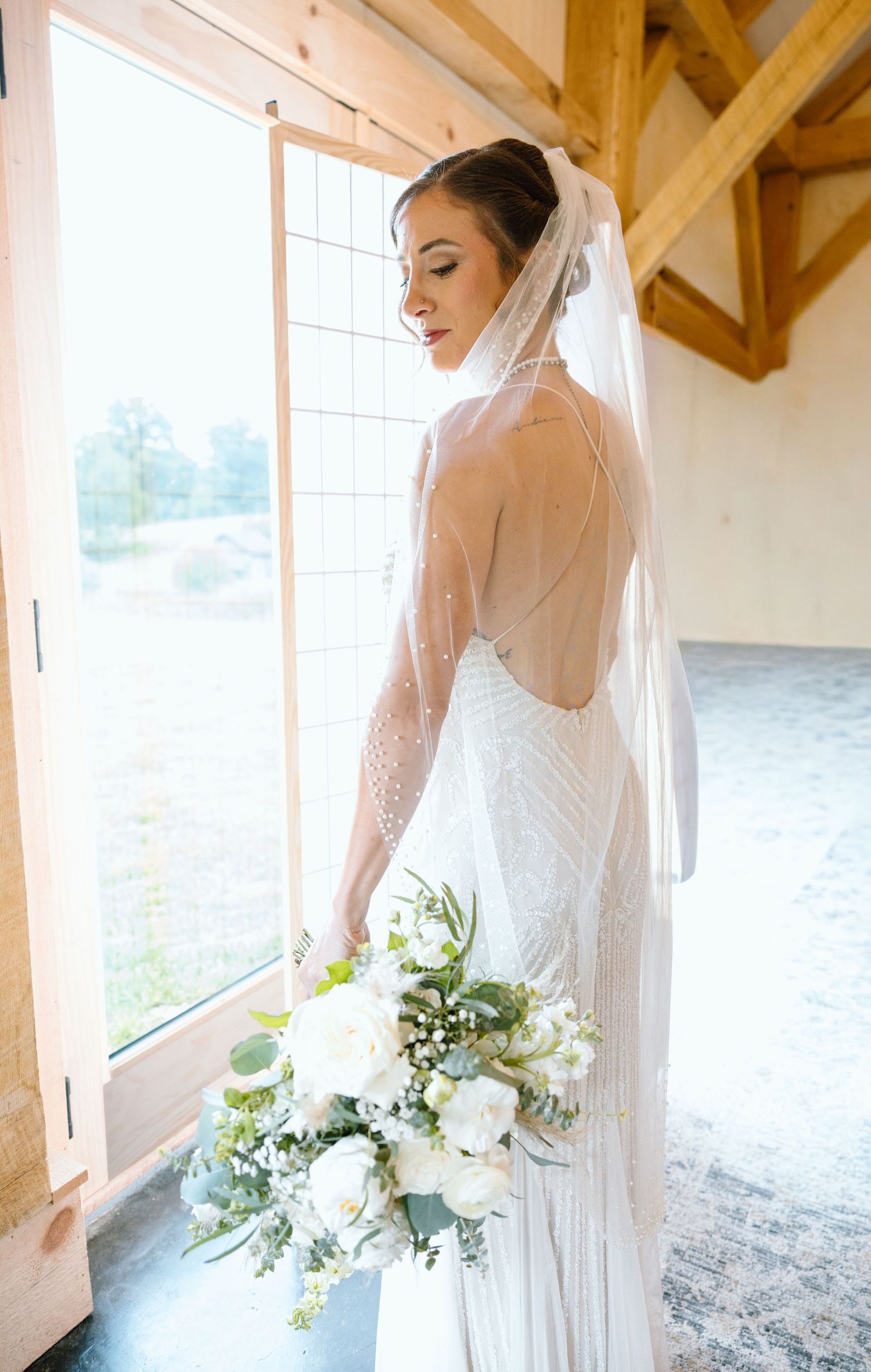 Bride in backless beaded gown and veil holds white floral bouquet by sunlit barn window.
