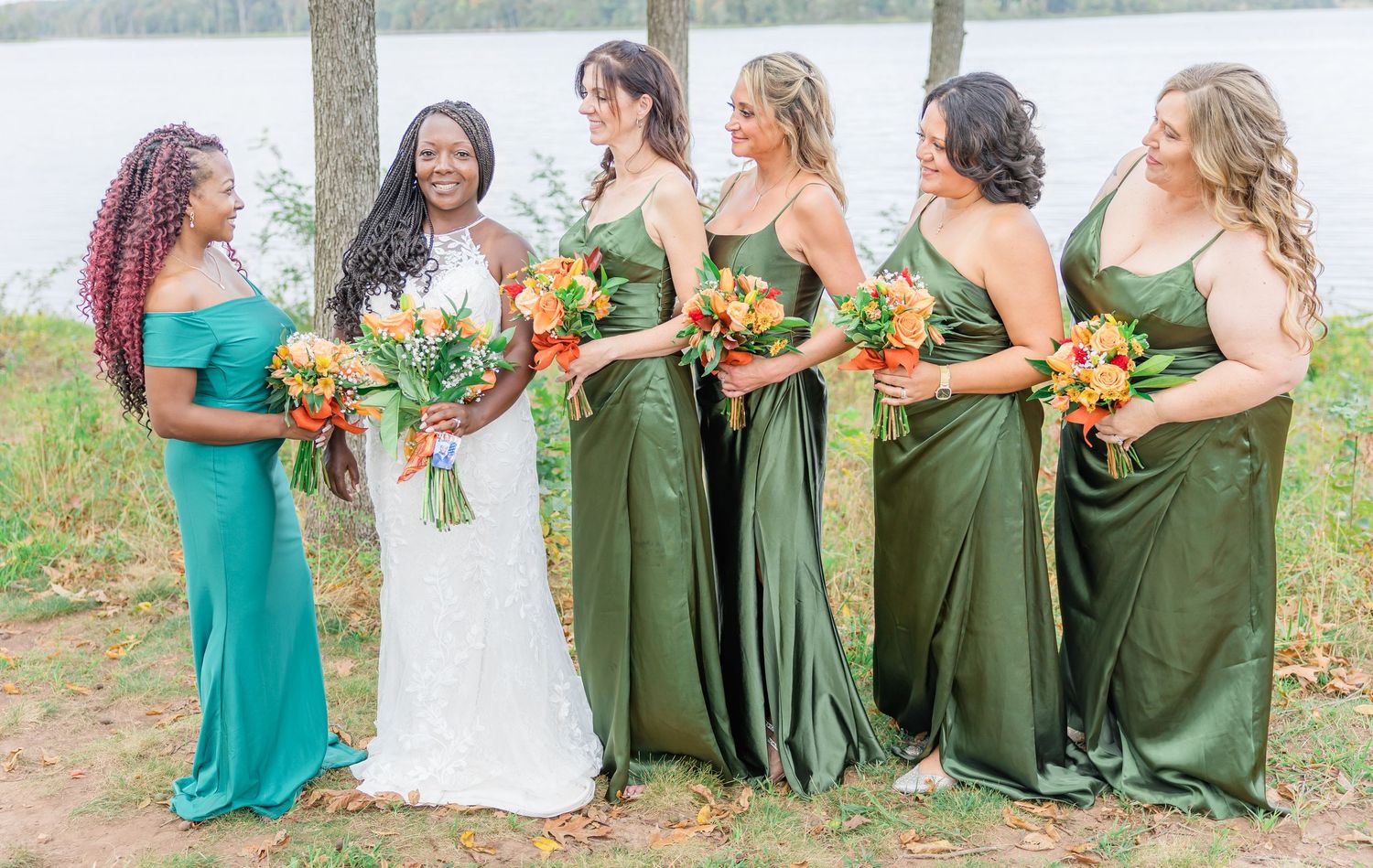 Row of bridesmaids in green dresses hold orange and white bouquets by the lake.