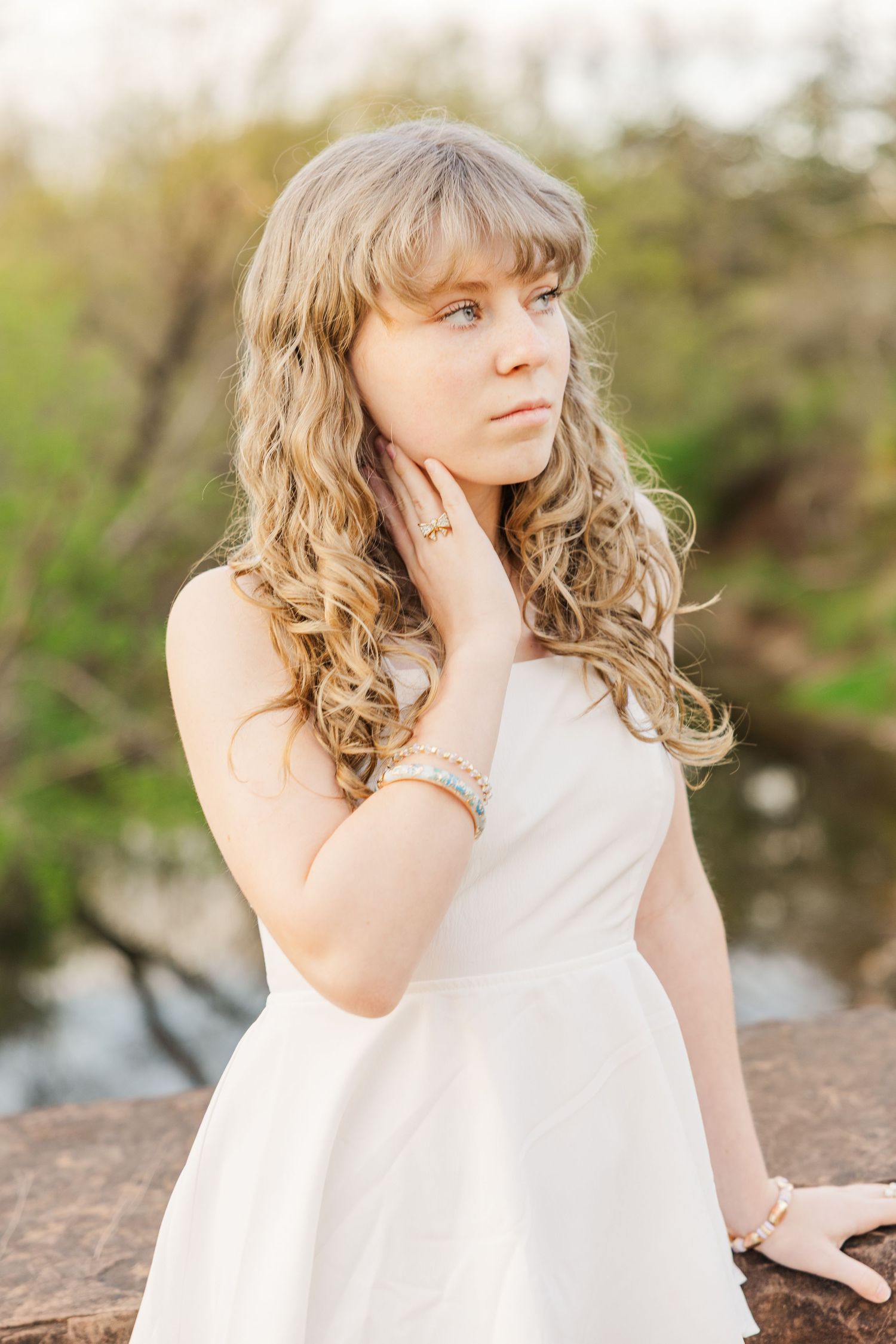 Graduate in white dress poses on rocky outcrop near water.