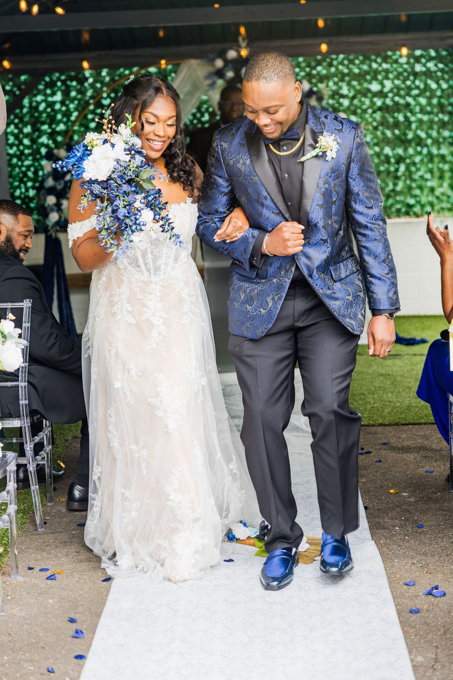 A couple in wedding attire laughs while performing a traditional jumping the broom ceremony at their outdoor reception.