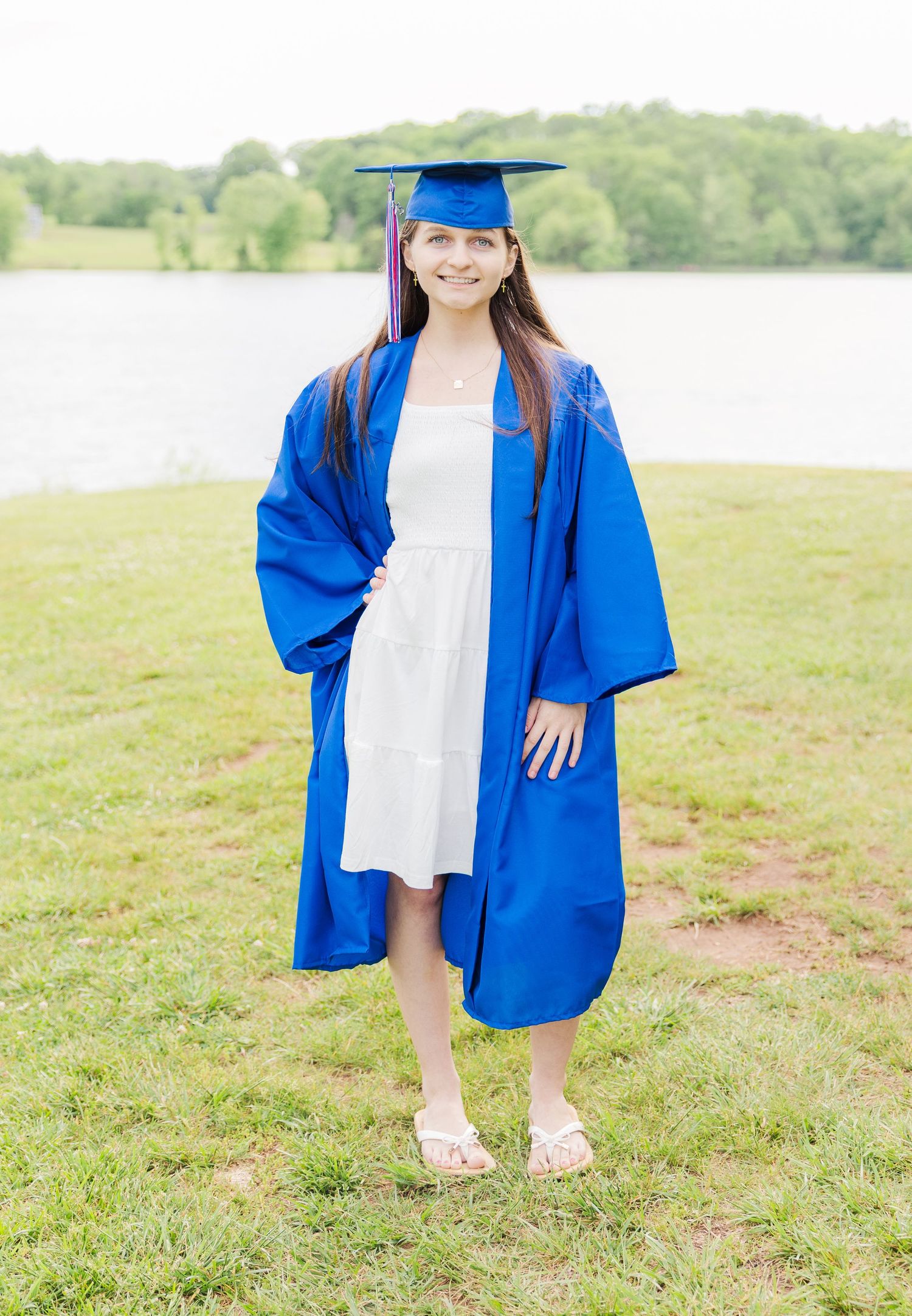 Child in blue graduation gown and white dress posing in grassy field by water.