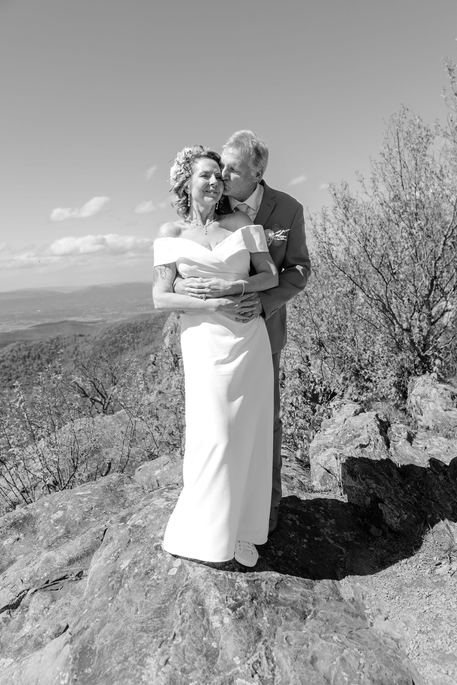 Black and white photo of a couple in wedding attire embracing on a rocky overlook with scenic mountain views.
