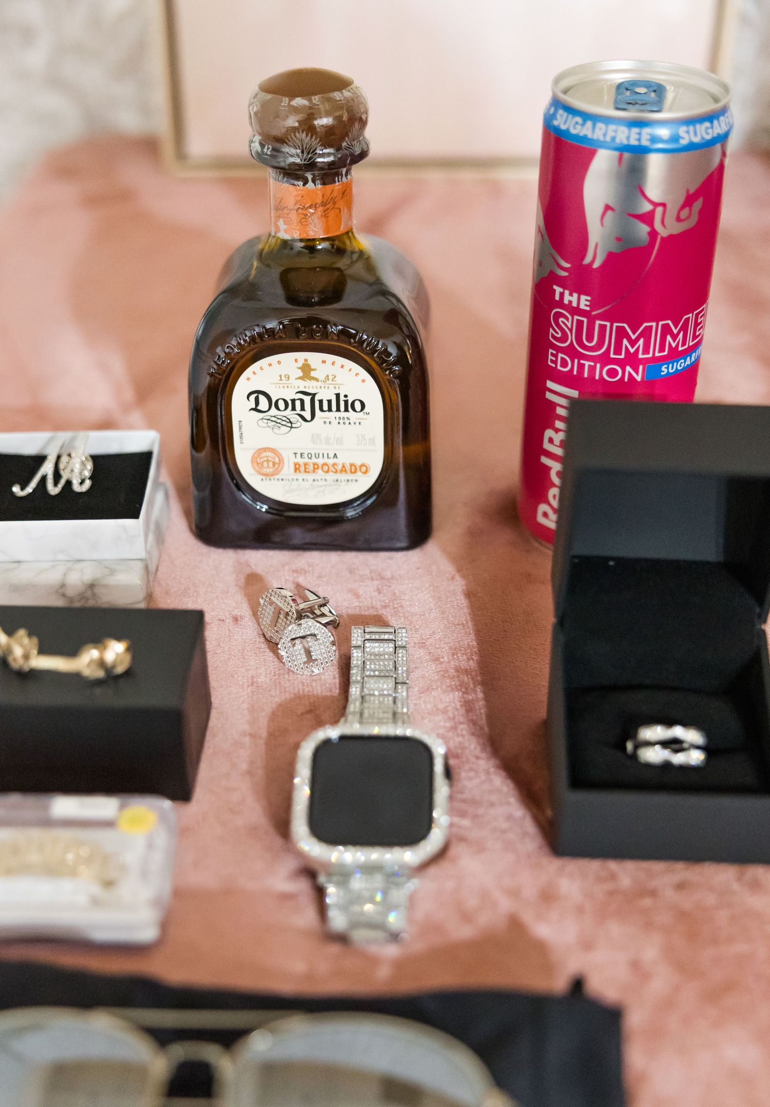 Wedding details including rings, jewelry boxes and bottles displayed on pink surface.