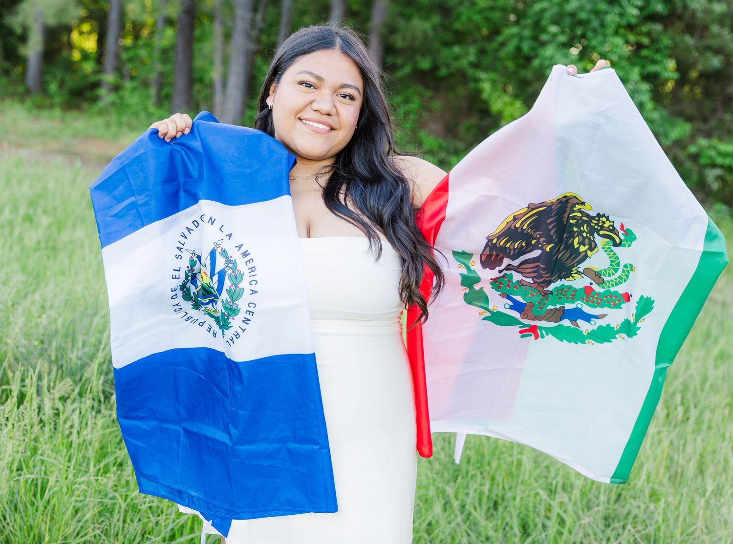 Person in white dress holding various Central American flags in a grassy outdoor setting.
