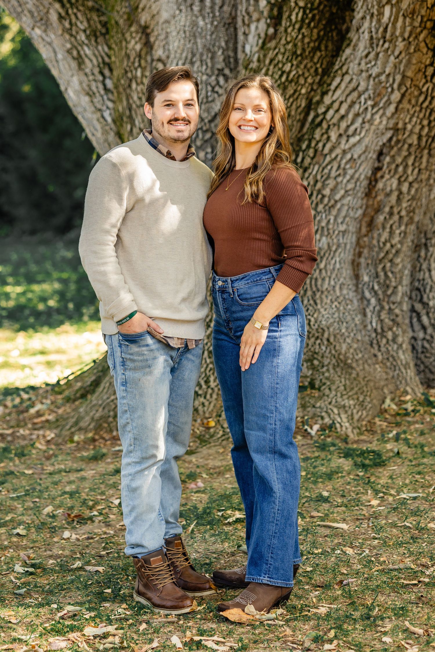 Couple in denim and neutral colors posing casually by large tree.