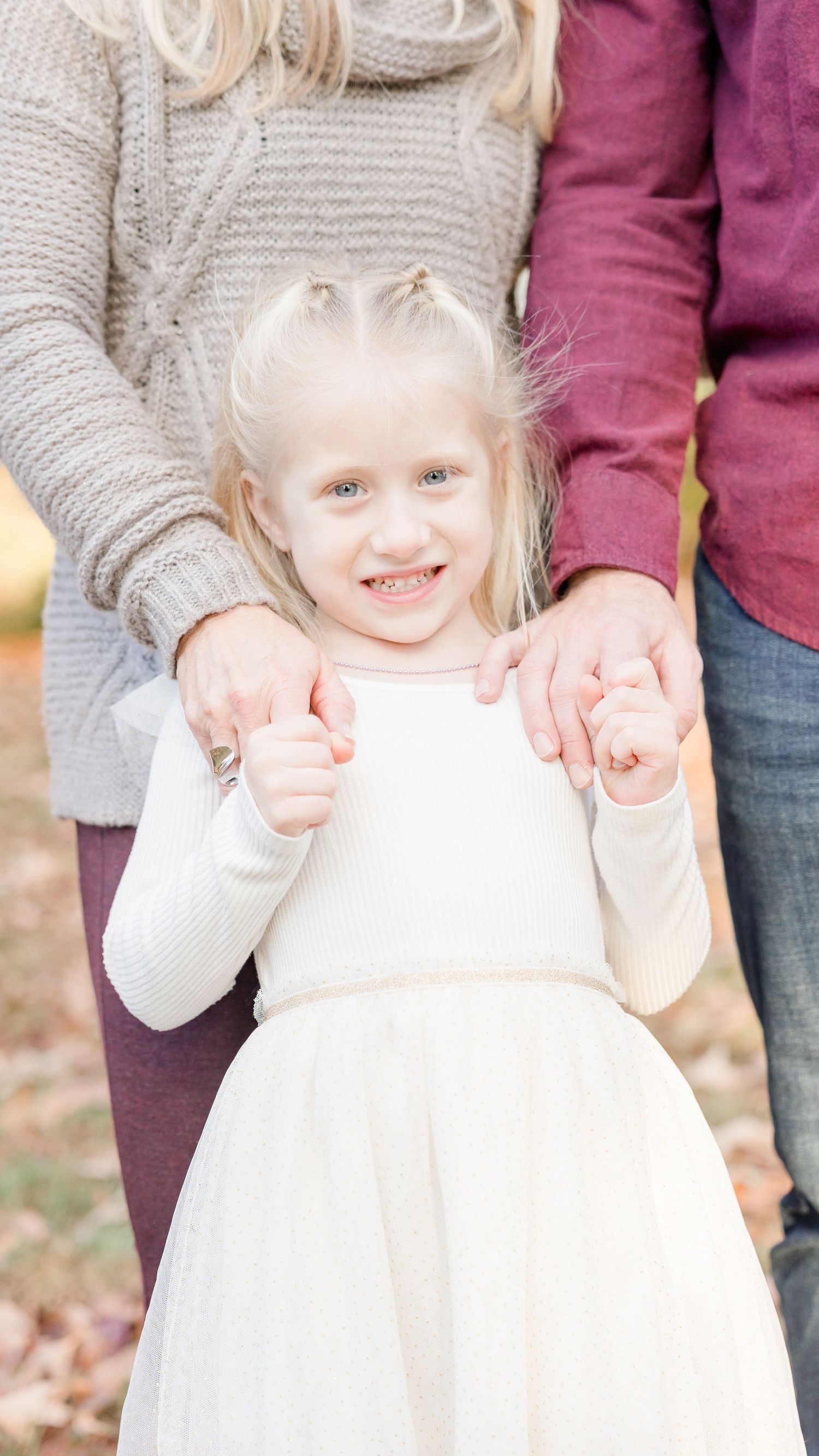 A young child in a white dress and gray cardigan smiles while being held by someone in burgundy and gray clothing.