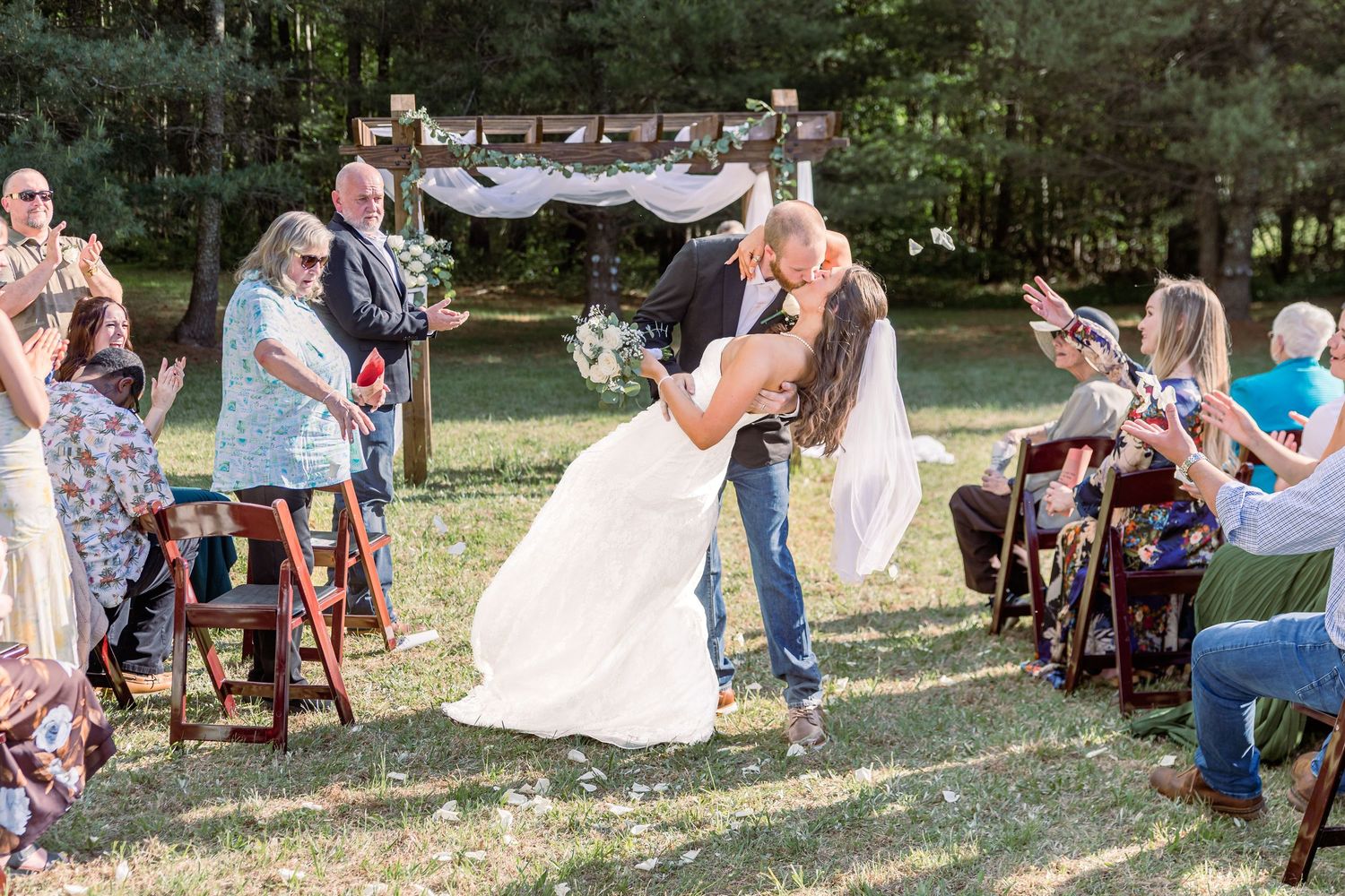 Bride and groom share a romantic dip kiss at outdoor wedding ceremony with guests cheering in rustic woodland setting.
