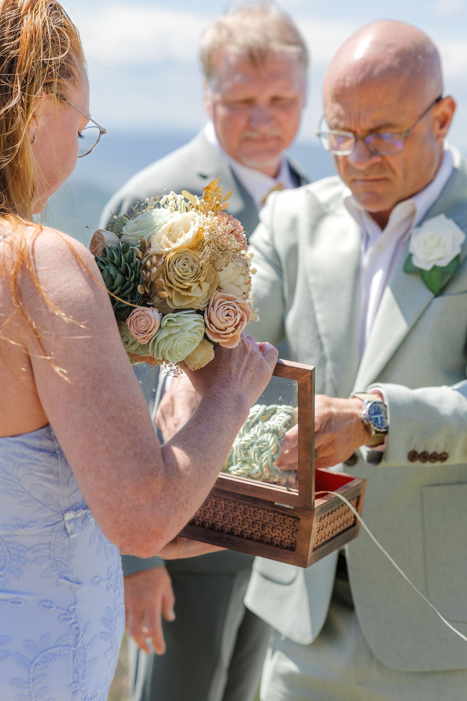Wedding ceremony with bride holding floral bouquet as groom opens wooden ring box, officiant standing behind them outdoors.