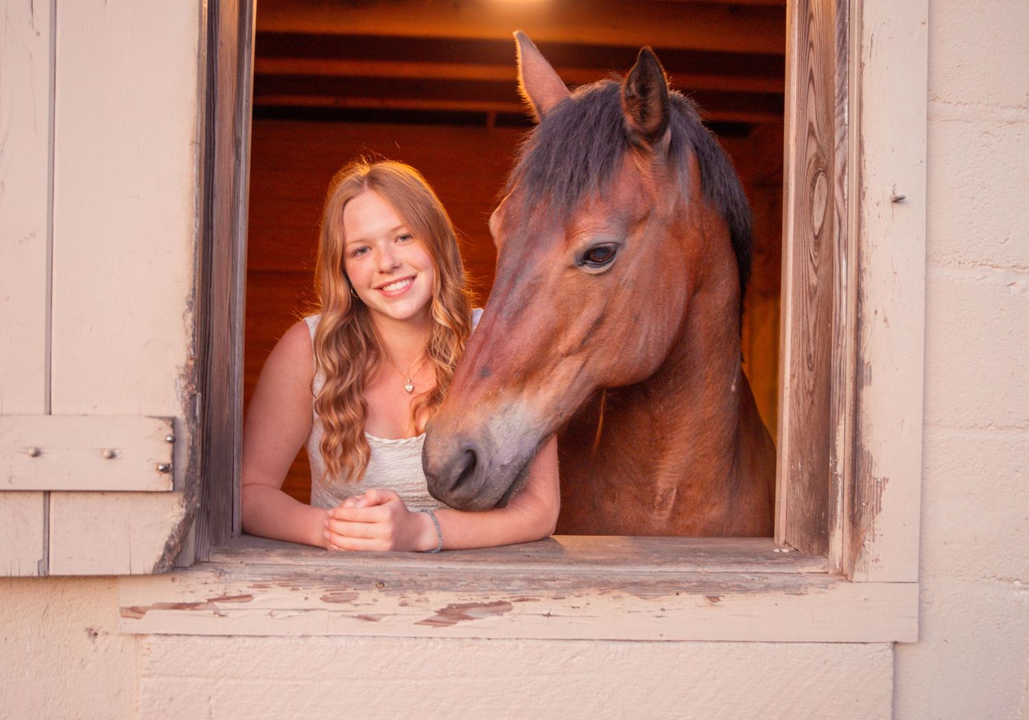 Smiling teenage girl with long blonde hair posing with a brown horse in a stable window opening.