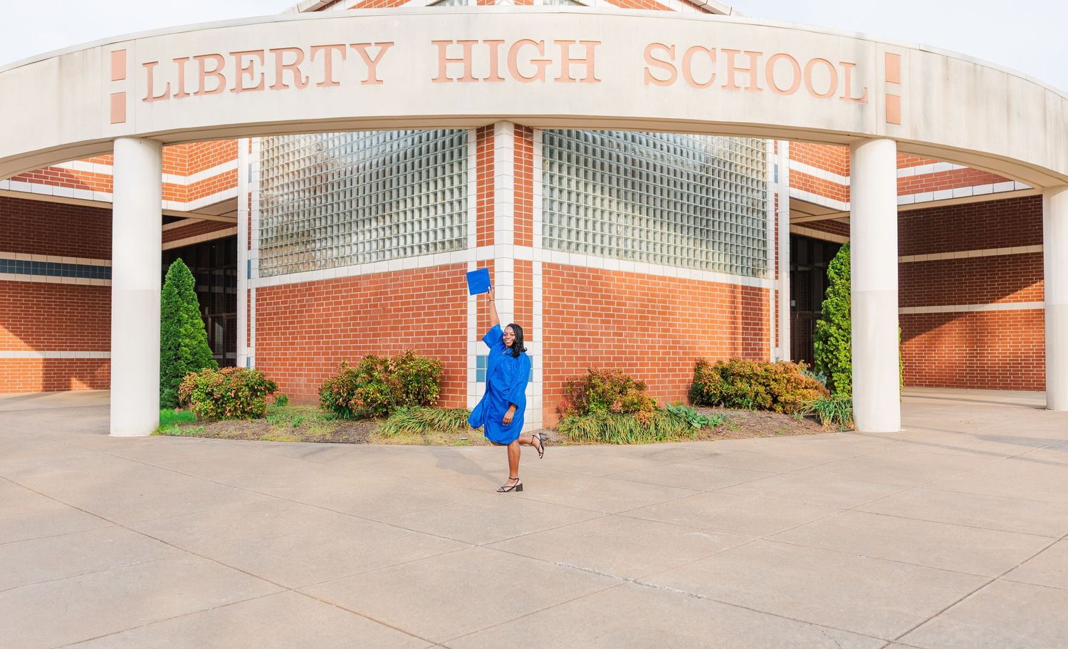 Graduate in blue cap and gown jumping for joy in front of Liberty High School entrance.
