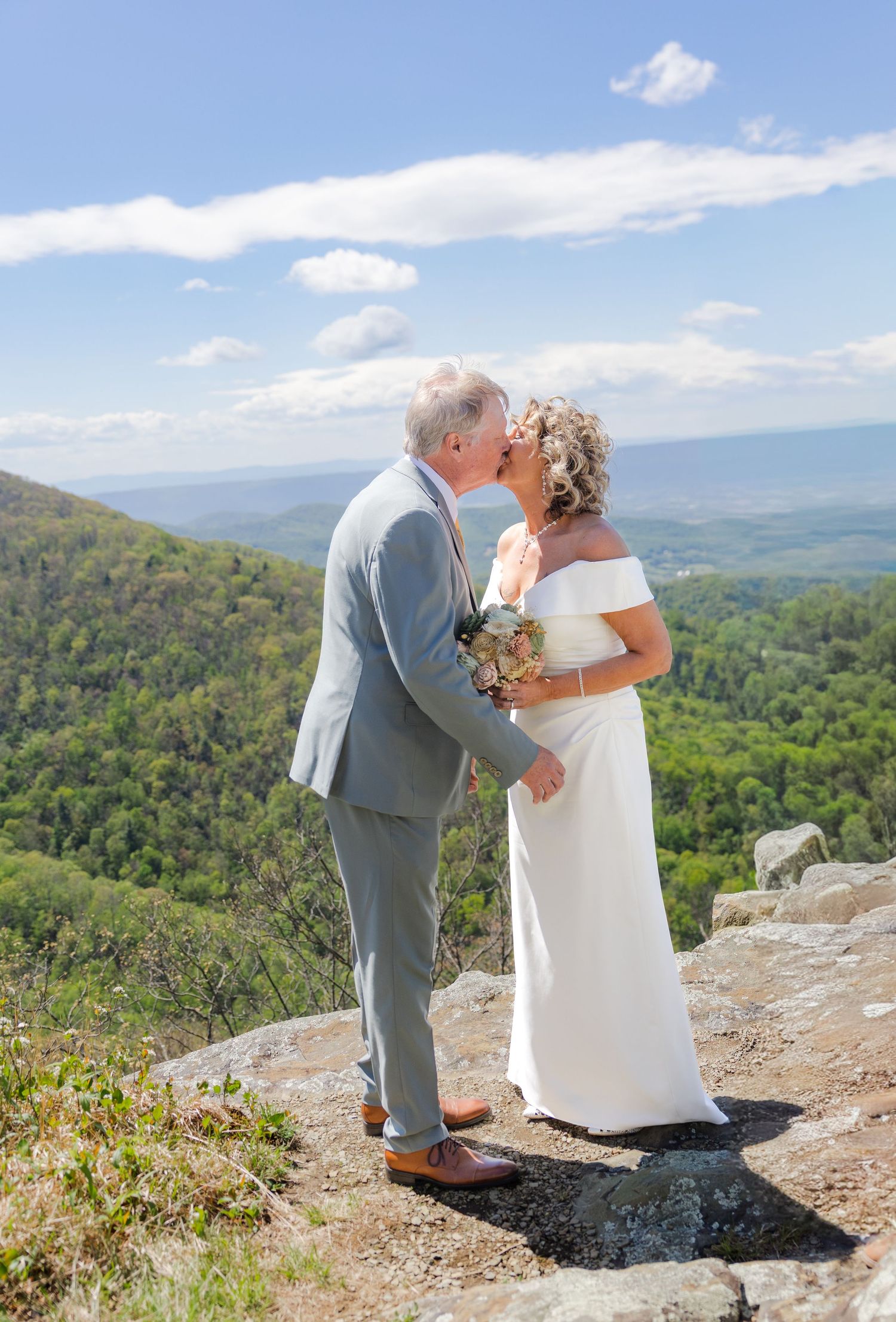 Couple sharing a kiss on a mountain overlook in wedding attire, bride in white gown, groom in gray suit, scenic valley view.