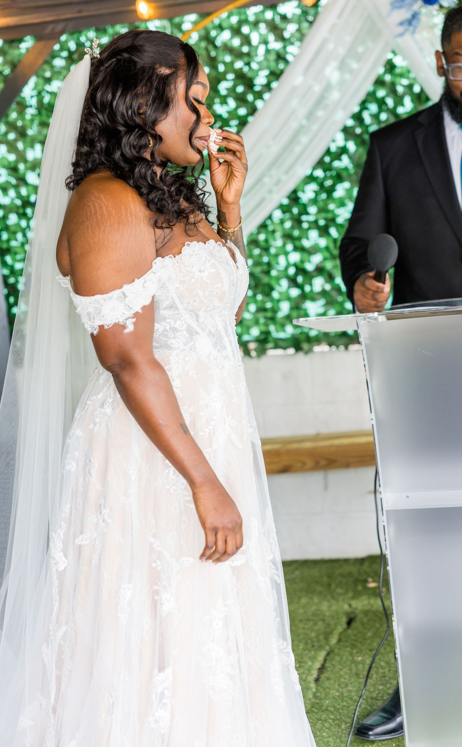 A bride in a flowing white lace wedding dress with off-shoulder detail stands in an elegant outdoor ceremony setting.