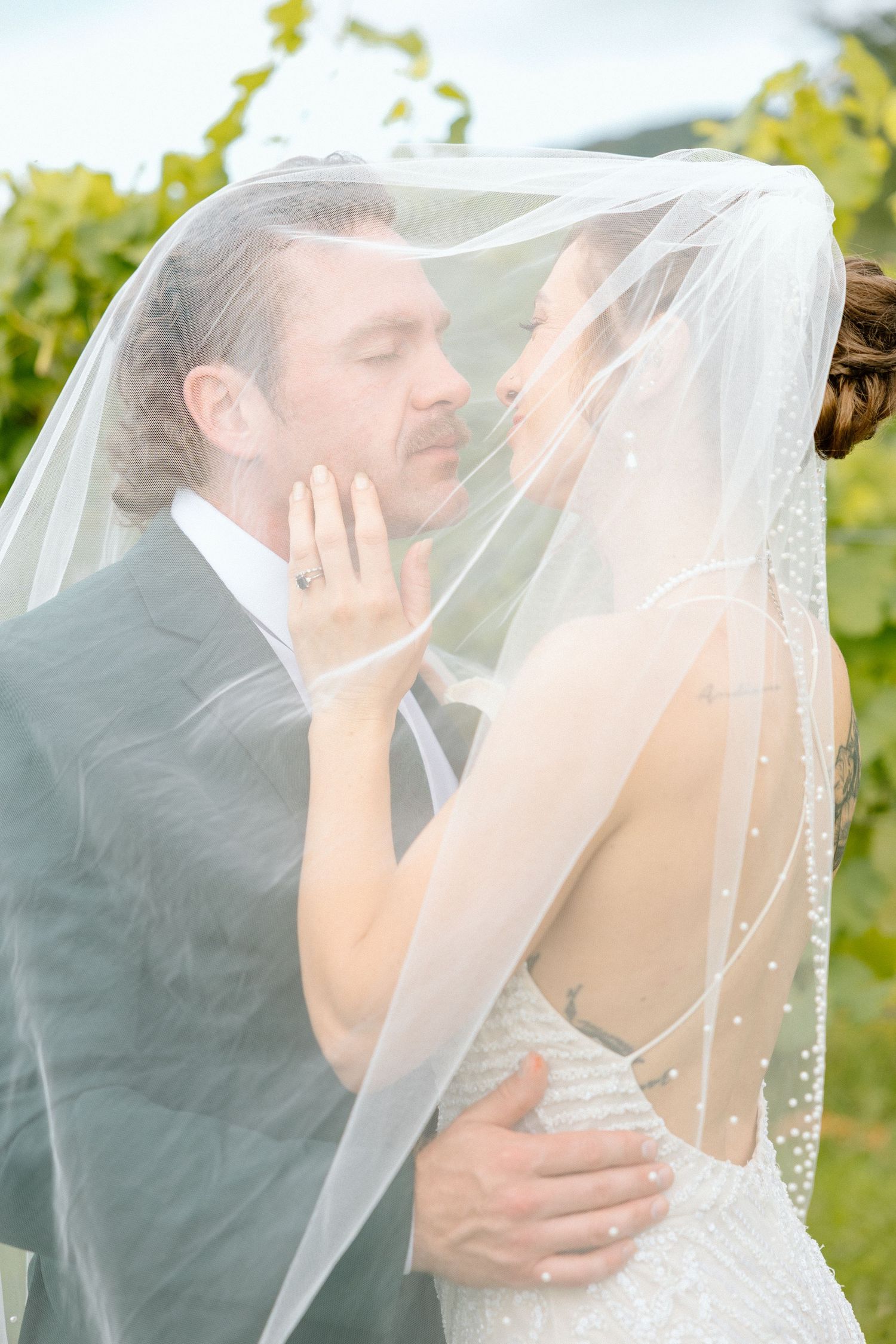 Bride and groom share a romantic moment under a sheer pearl-adorned veil at a vineyard wedding.