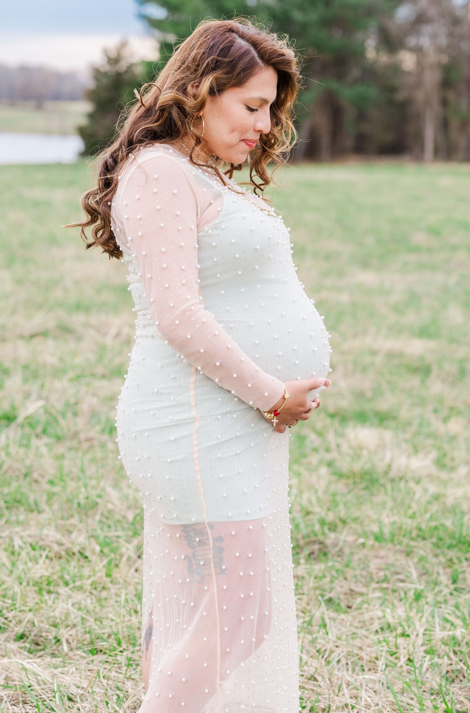 Maternity photos showing a woman in a delicate white dress with pearl details in a grassy field.