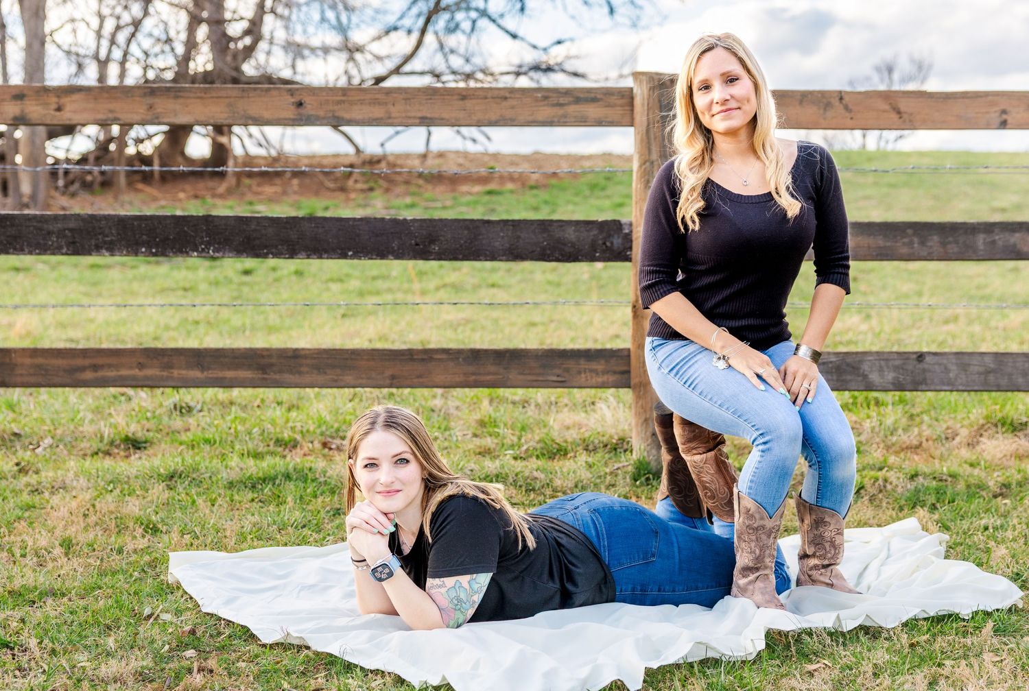 Two friends relaxing on a blanket by a wooden fence in a pastoral setting.