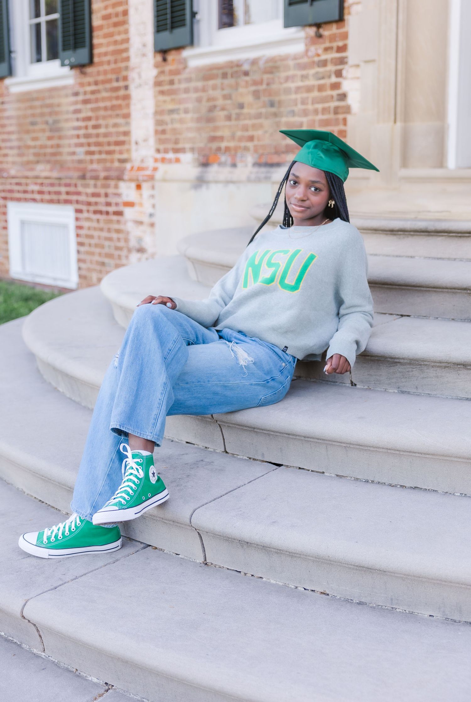 Graduate wearing green cap and NSU sweatshirt relaxes on concrete steps.