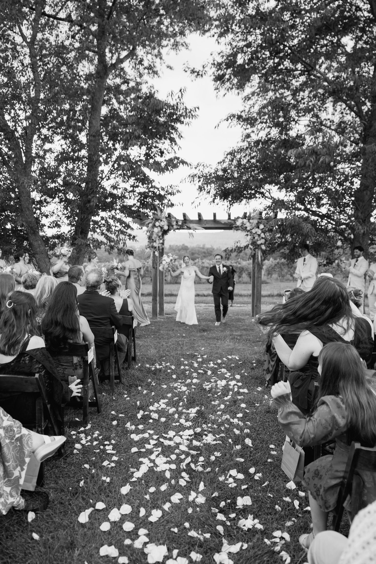 Black and white outdoor wedding ceremony with couple at floral arch, petal-lined aisle, and seated guests among trees.