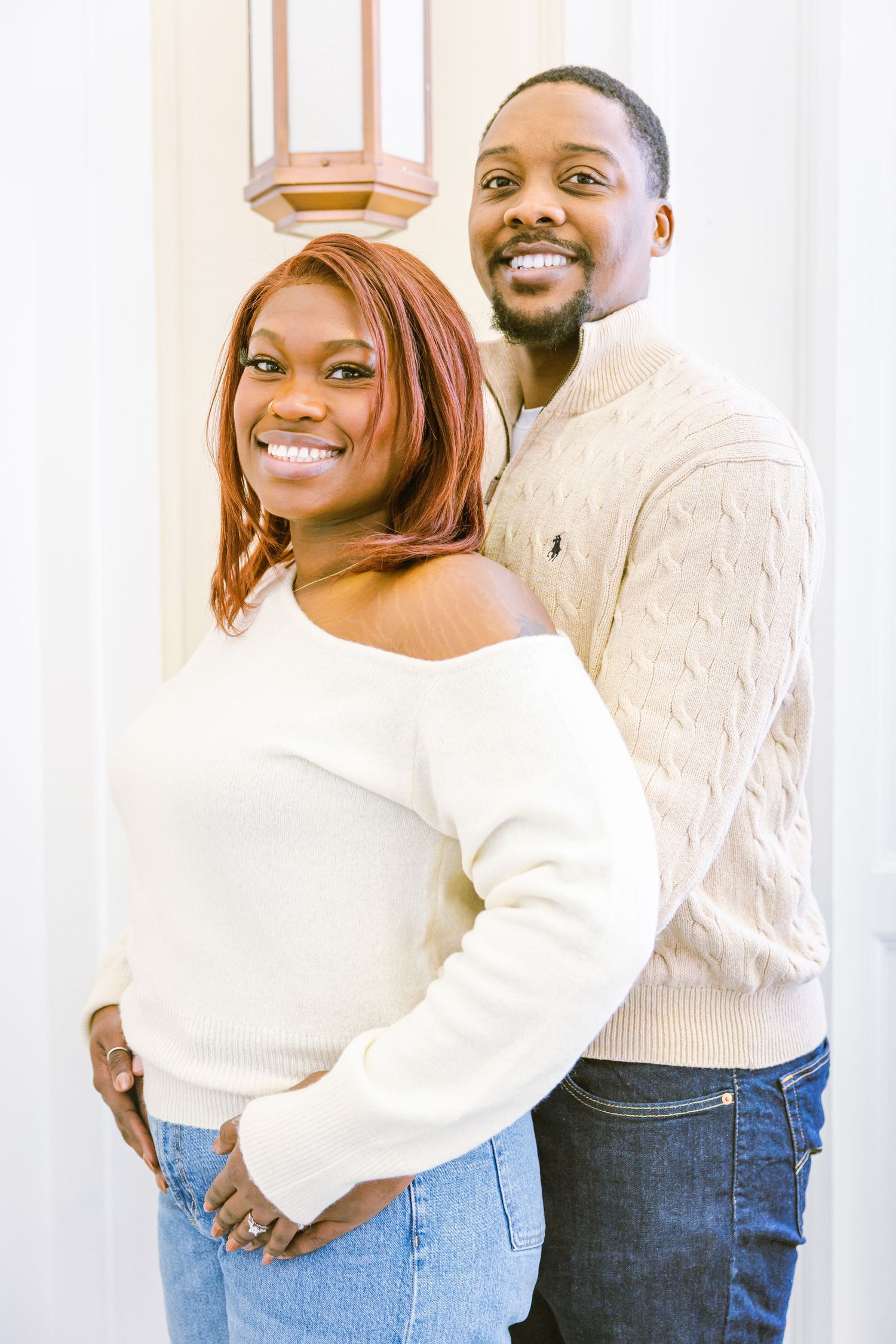 A couple wearing cozy white sweaters poses together against a light background, sharing warm smiles.