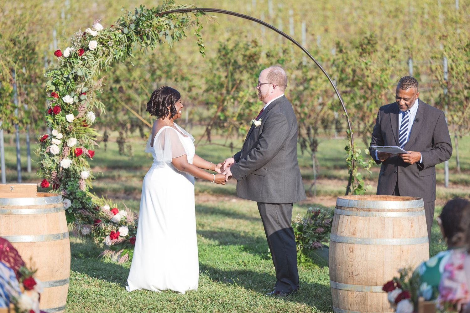 Outdoor vineyard wedding ceremony with wine barrels and floral decorations.