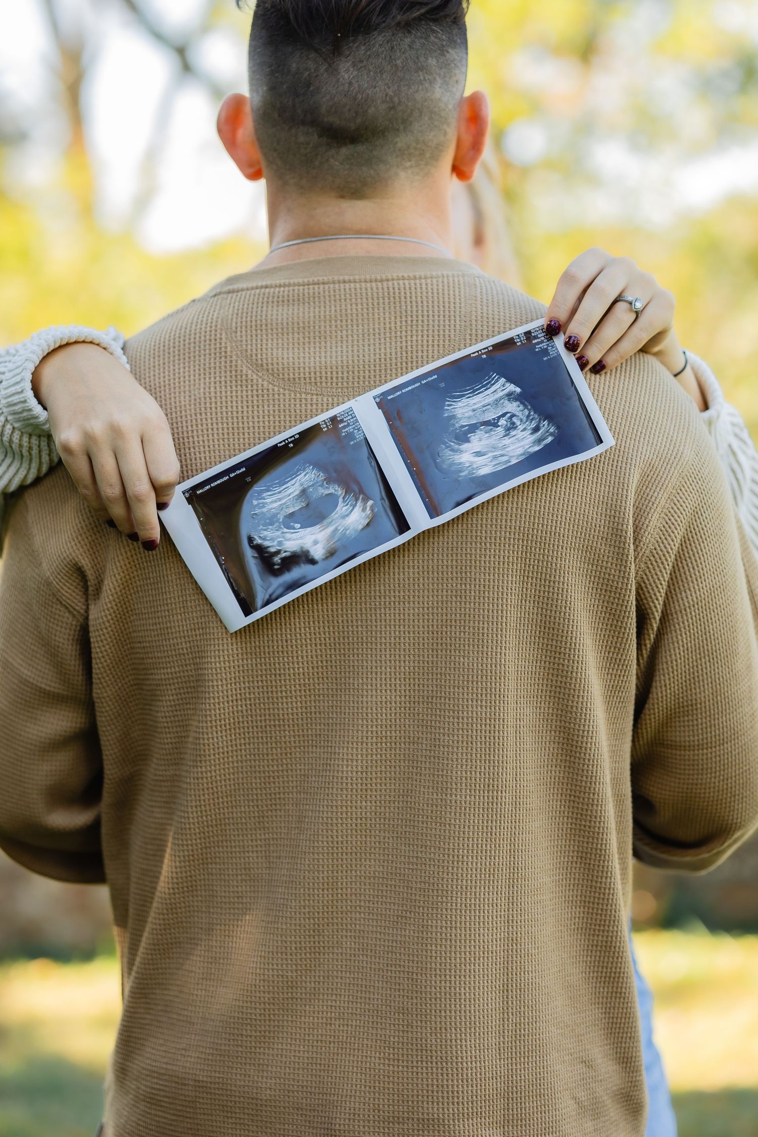 Back view of person holding ultrasound images against beige sweater in sunlit setting.