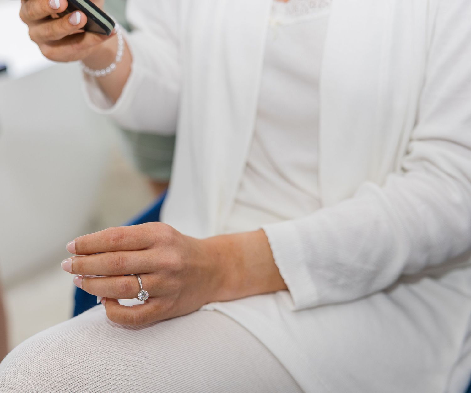 Woman in white outfit wearing a diamond ring, holding a smartphone while seated.