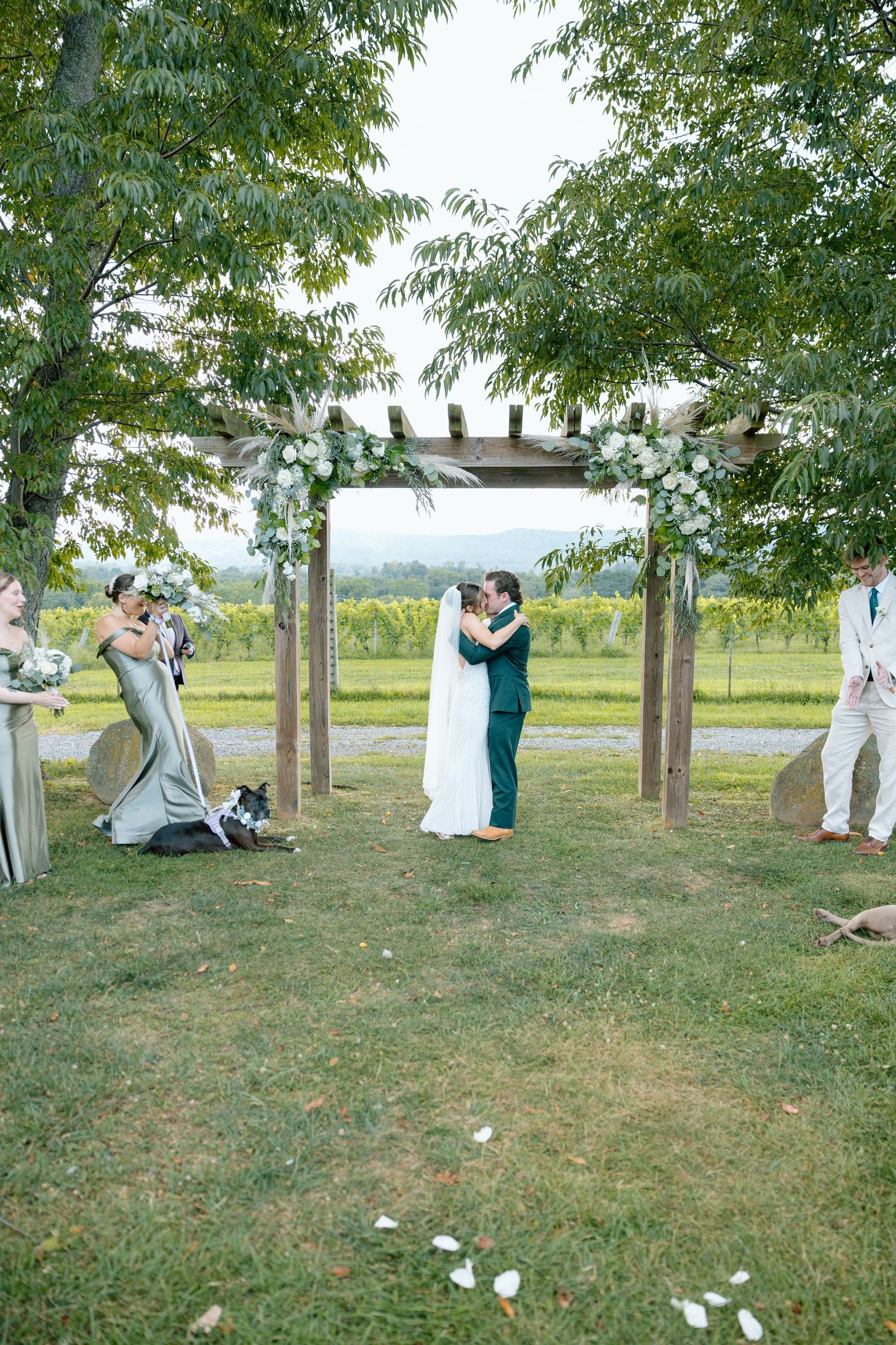Couple shares first kiss under floral pergola arch at outdoor vineyard wedding ceremony with wedding party and dog.