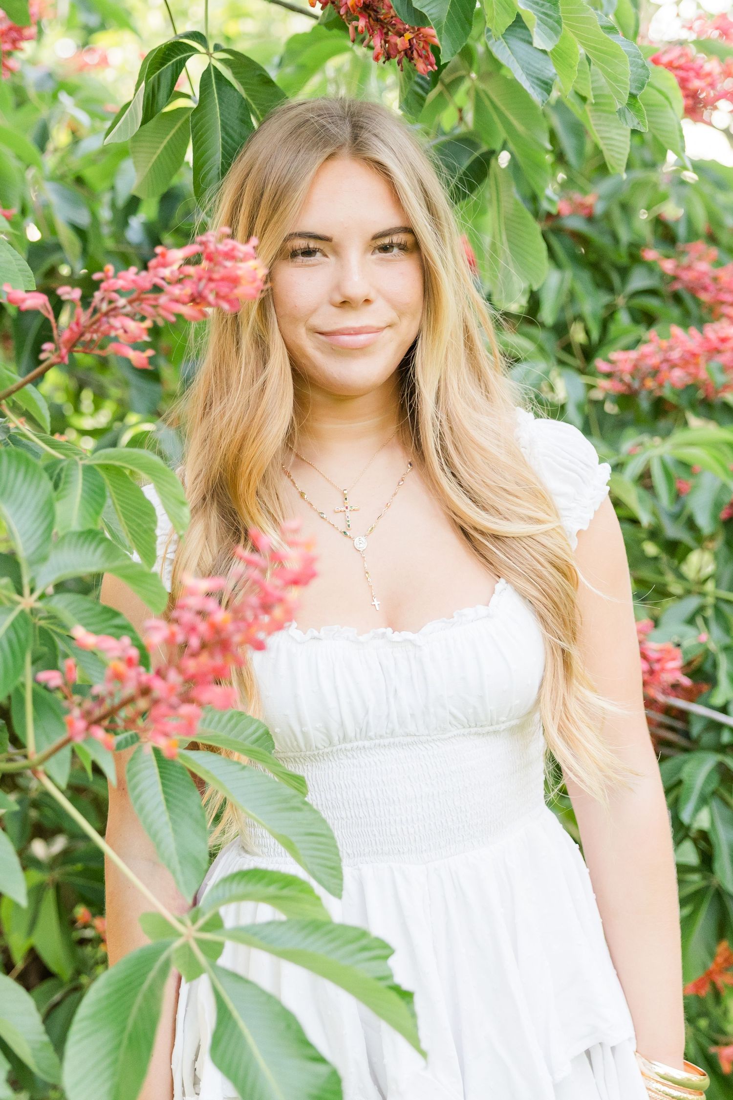 Woman in white summer dress posing near flowering bushes with red blooms.