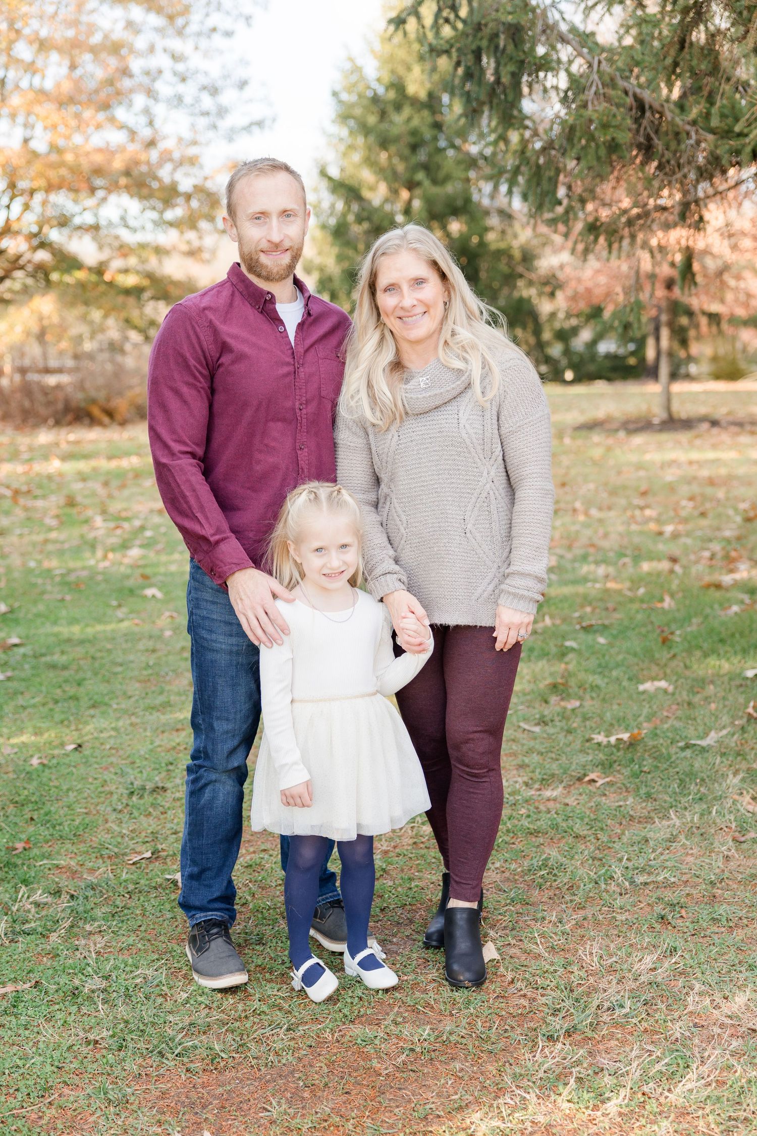 Family in coordinating burgundy and beige outfits poses together on grass during autumn photoshoot.