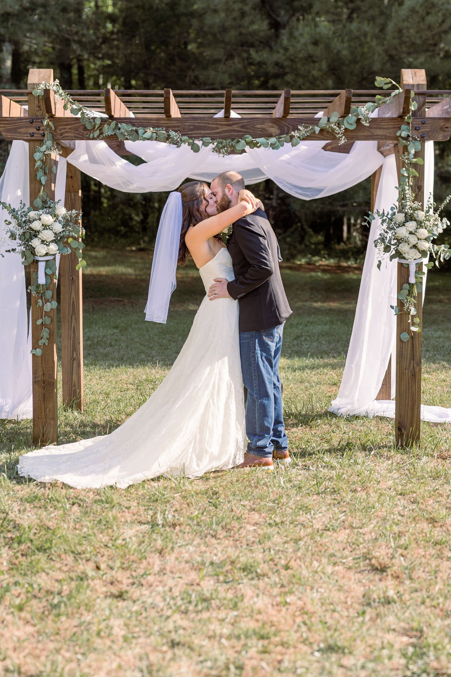Bride and groom share first kiss under rustic wooden arch with white draping and floral arrangements outdoors.