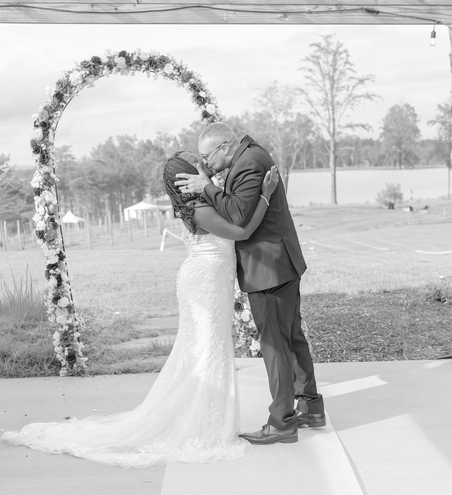 Black and white photo of newlyweds sharing their first kiss under a decorated arch.
