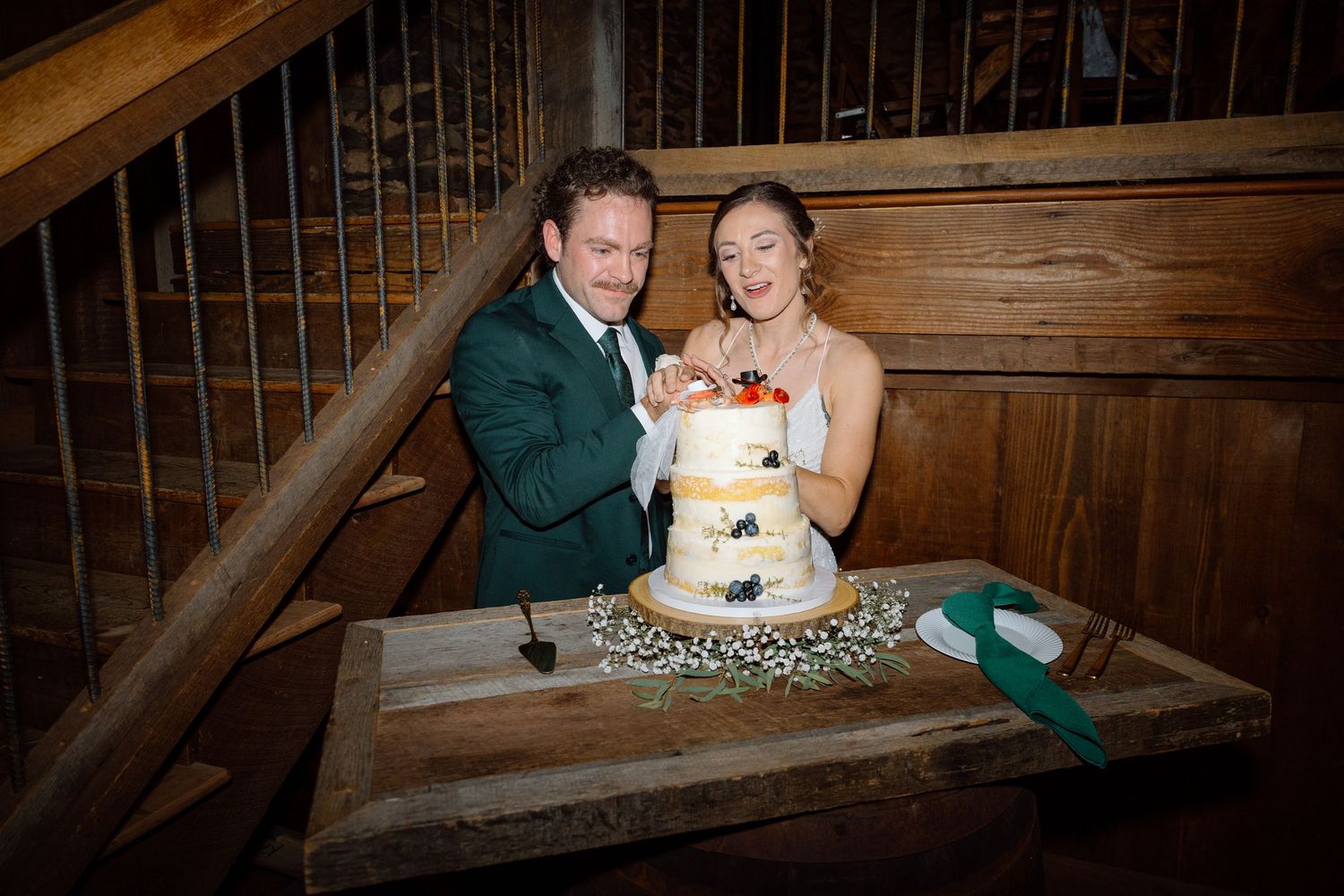 Bride and groom cutting a three-tier wedding cake decorated with fresh fruit at a rustic barn venue.