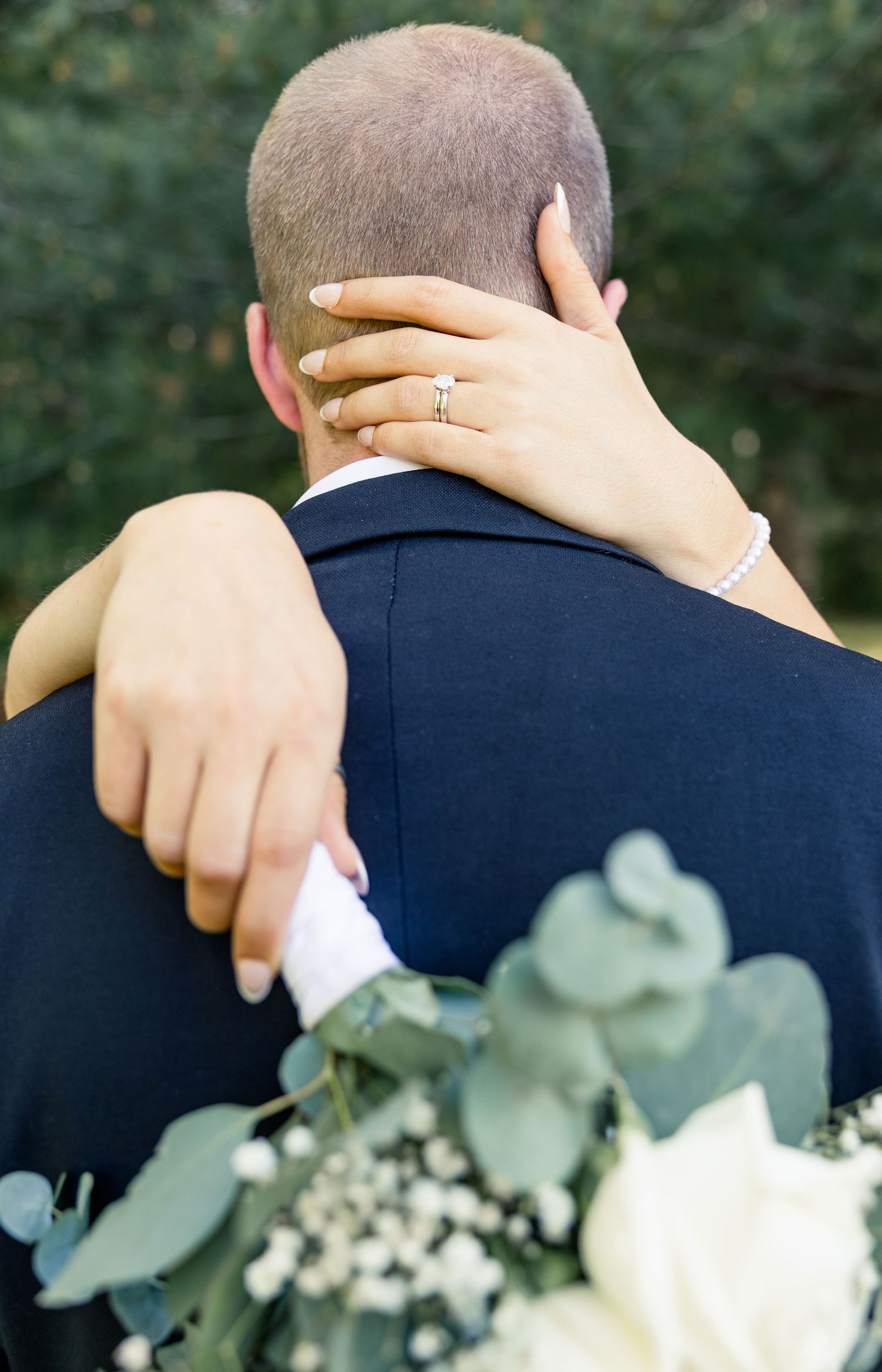 Bride resting hand on grooms shoulder showing wedding ring while holding white floral bouquet with eucalyptus.