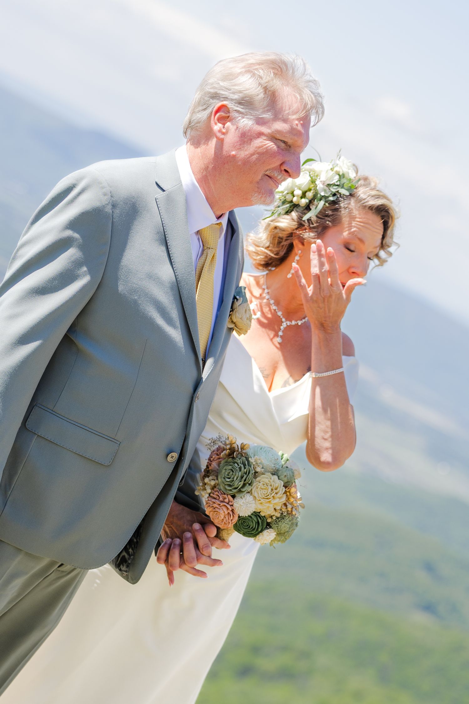 Bride and groom share emotional moment outdoors with mountain views, bride holds succulent bouquet wearing floral crown.