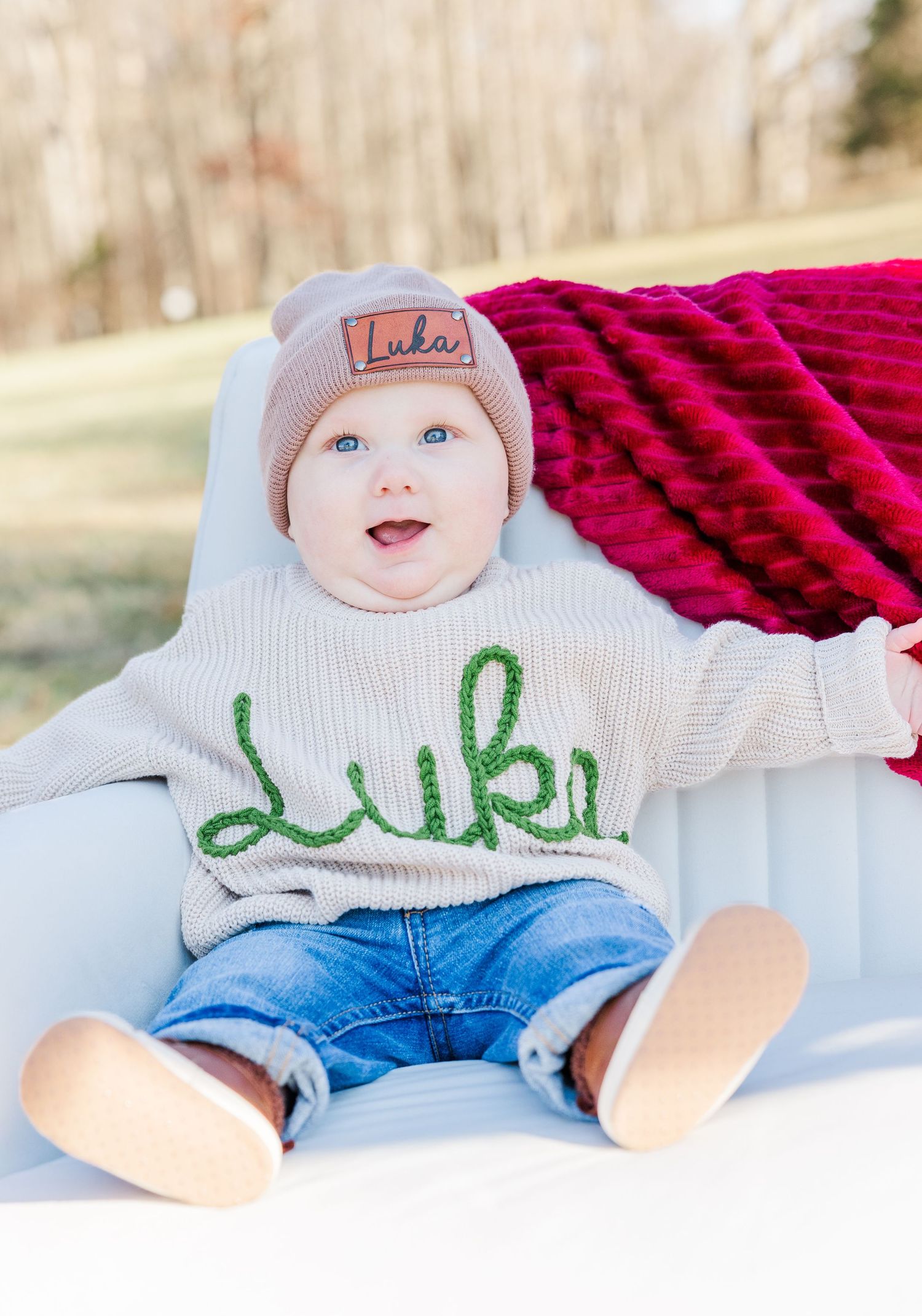 Baby wearing white sweater and jeans lies on red blanket outdoors.