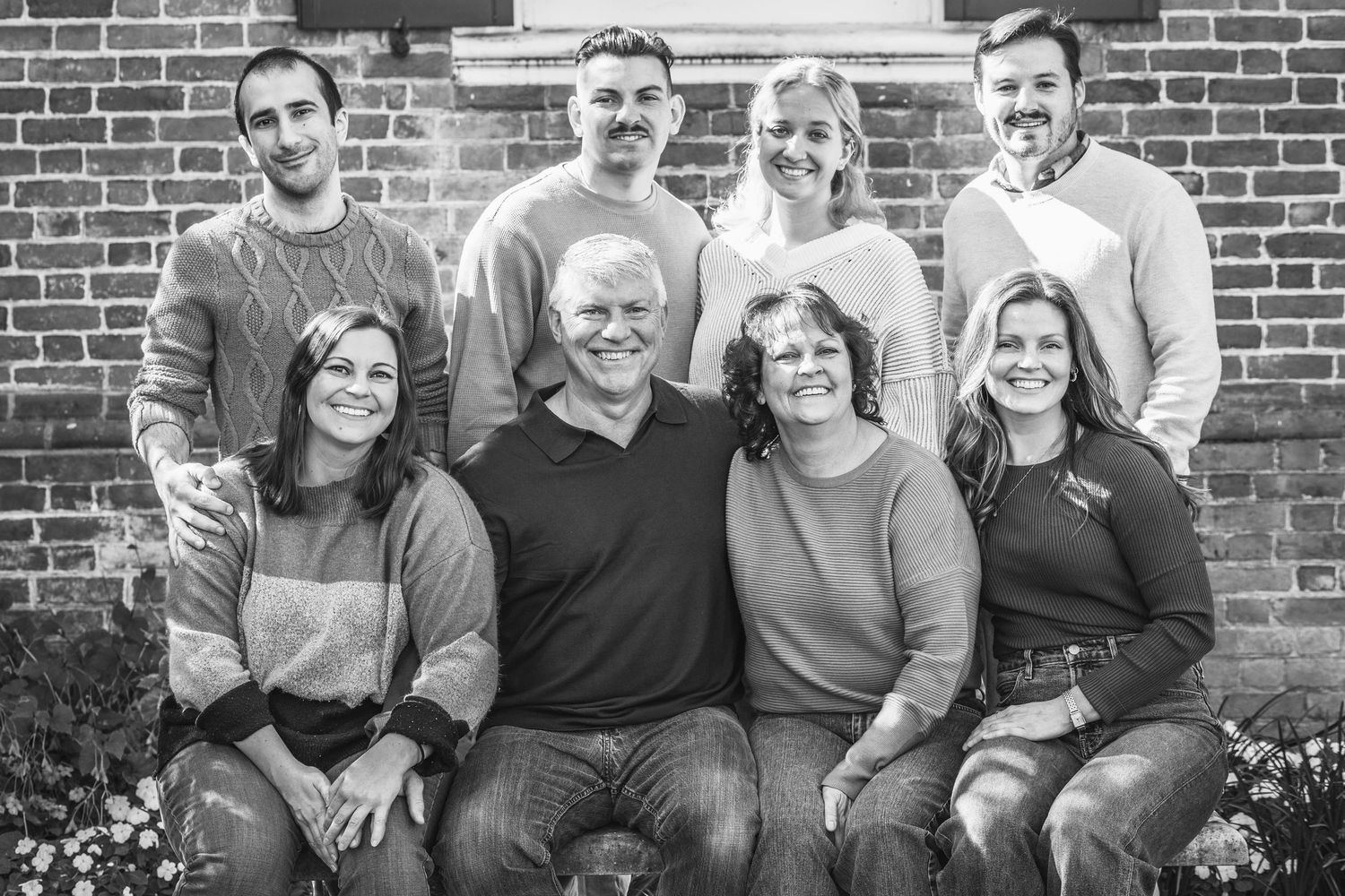 A black and white family portrait of eight people arranged in two rows against a brick wall.