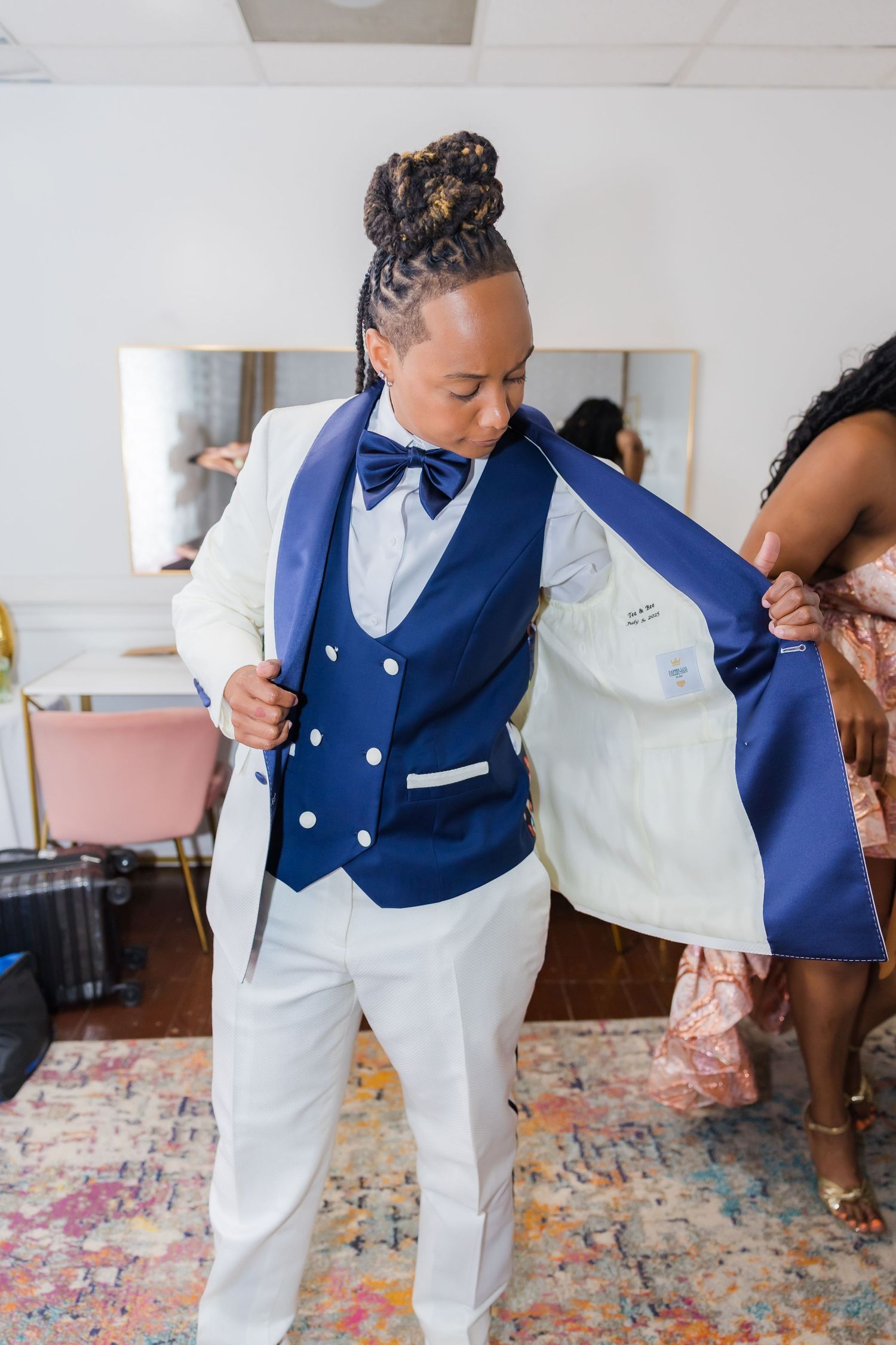 Someone in a white suit and navy vest bending down on a colorful rug.