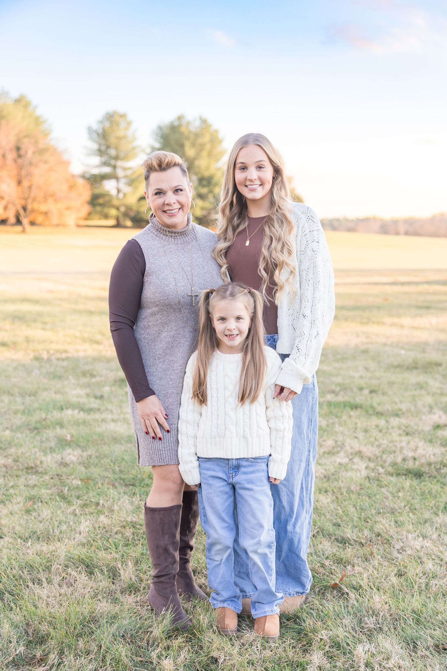 Three people stand together in a grassy field during autumn wearing cozy sweaters and casual attire.