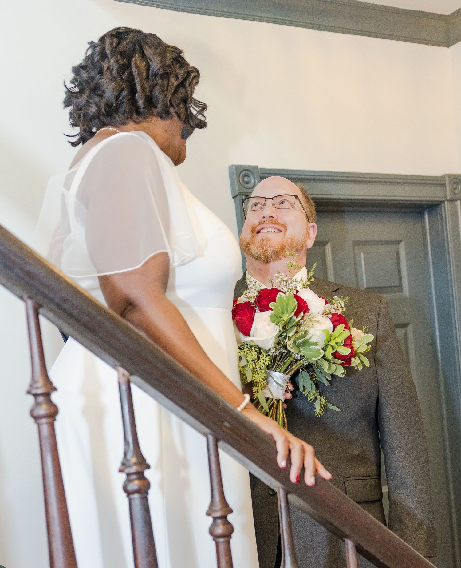 Couple sharing a moment on wooden staircase with red and white floral bouquet.