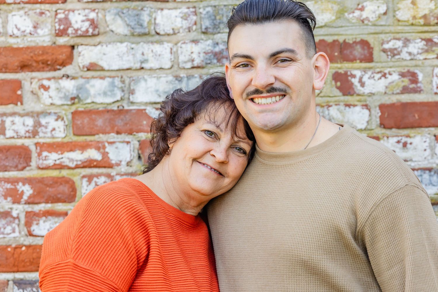 A couple poses together against a weathered brick wall, wearing orange and beige sweaters.