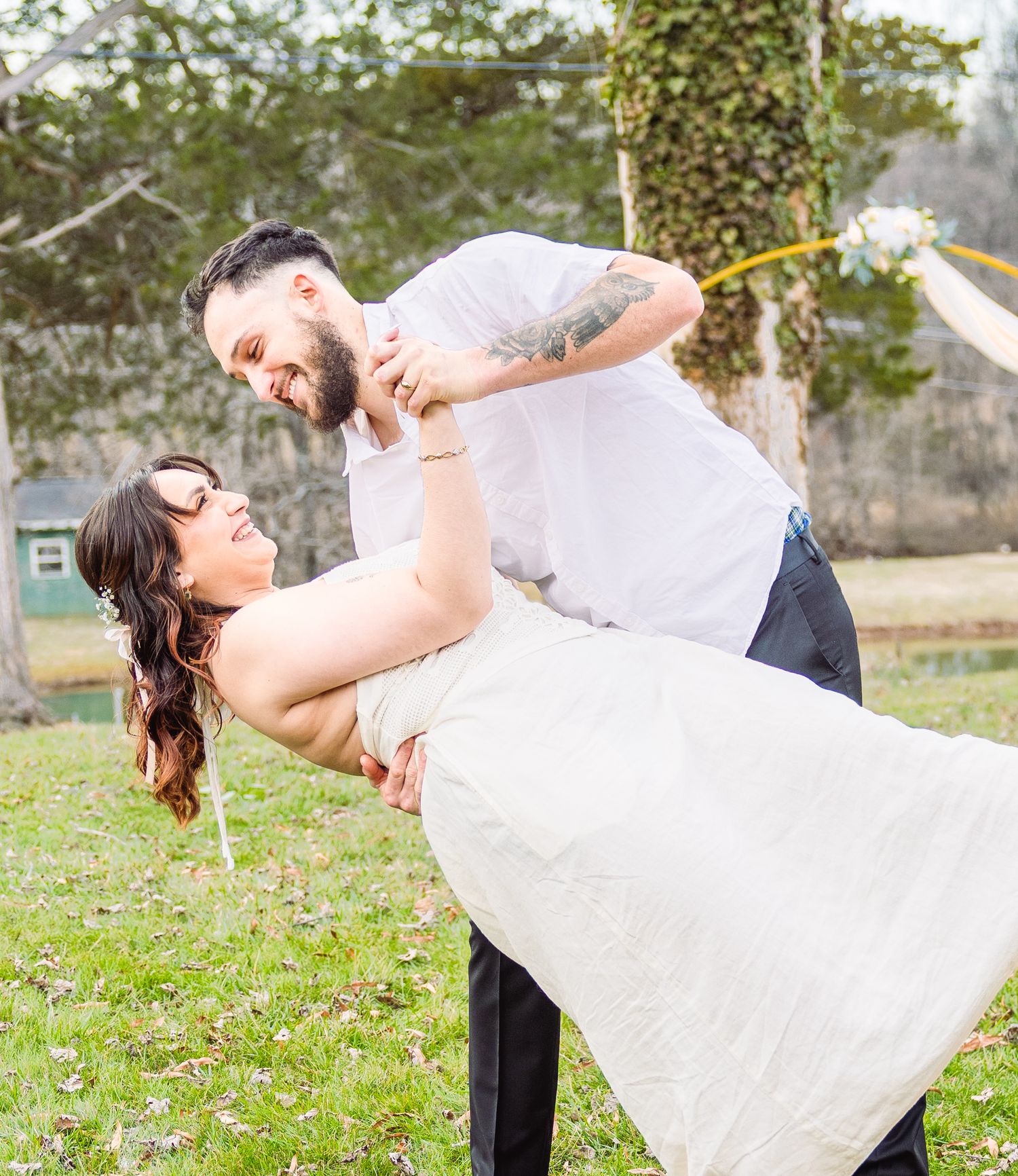 A couple in wedding attire performs a romantic dip pose together on grassy lawn during their outdoor photo session.