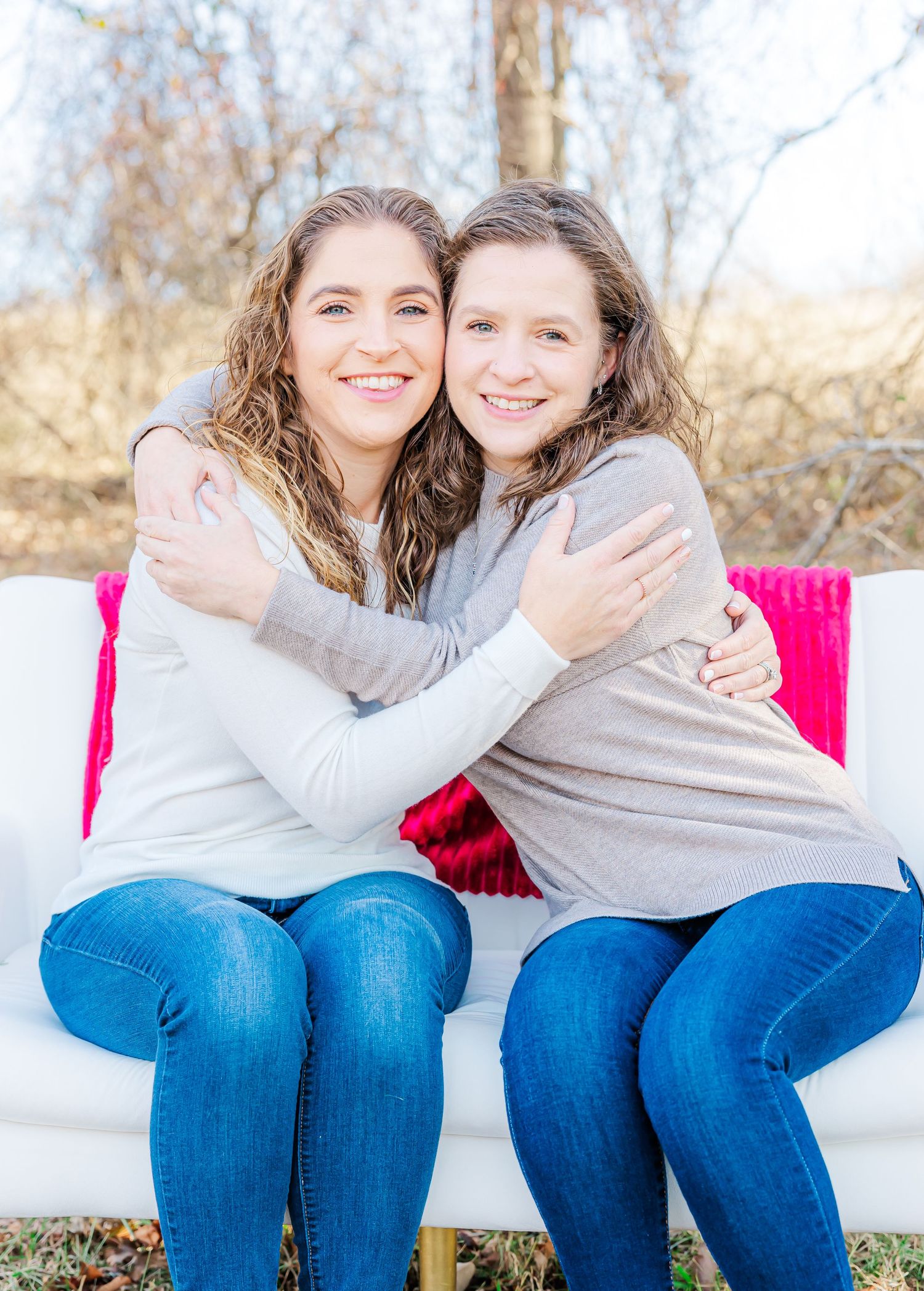 Two friends in matching sweaters and jeans share warm embraces on white bench.