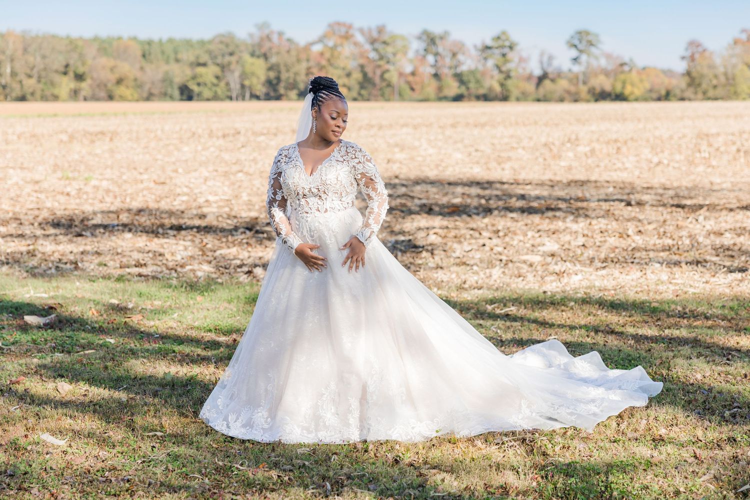 A bride in a flowing white gown sits gracefully in a sunlit autumn field during an outdoor bridal photoshoot.