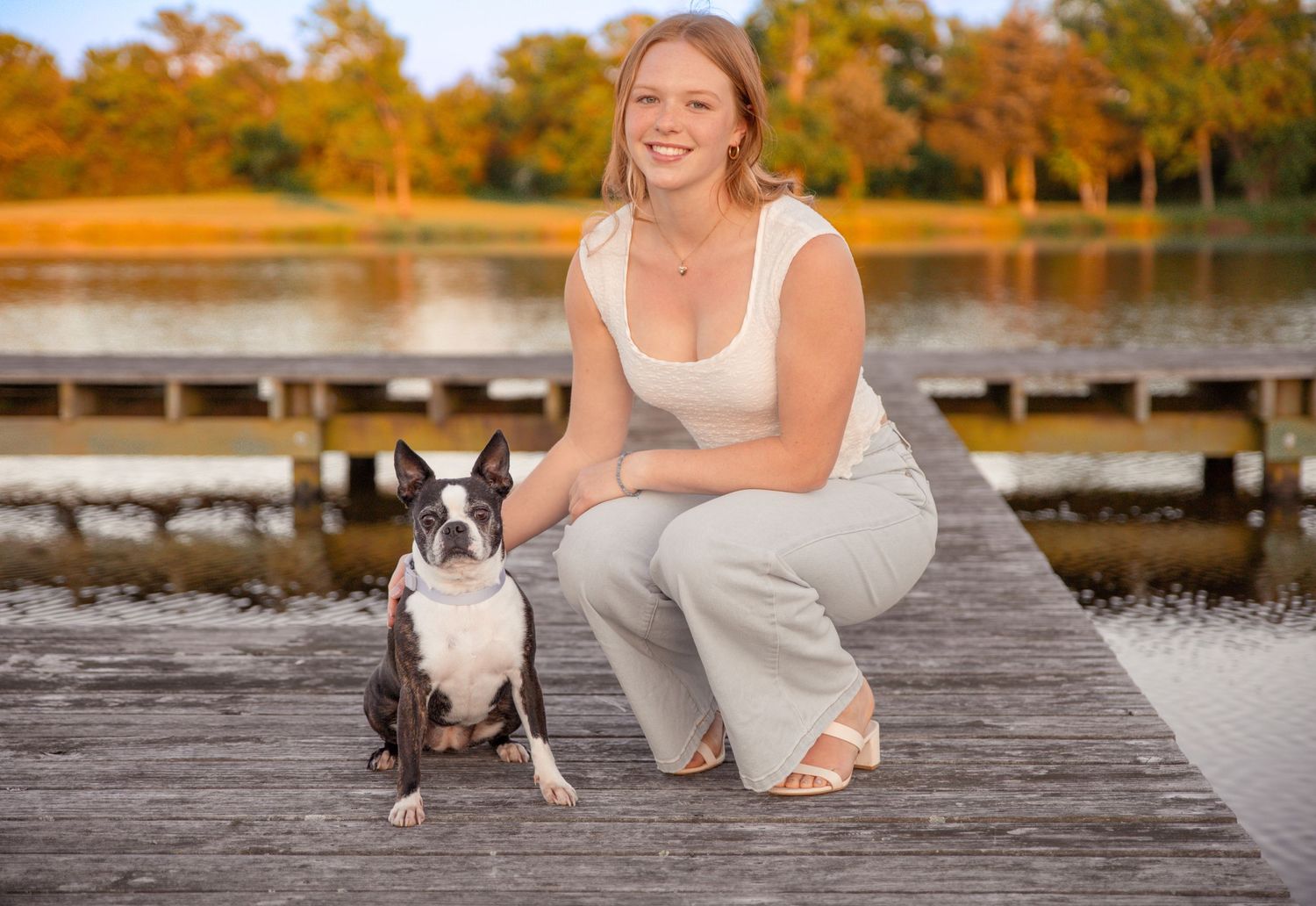 Young woman in white outfit poses with Boston Terrier dog on wooden dock by autumn lake.